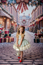 A young girl stands in a festive Christmas town street wearing a light green and cream tiered dress with red accents, holding a lantern. The background features decorated storefronts, Christmas trees, wrapped gifts, and a large red bow hanging from above.