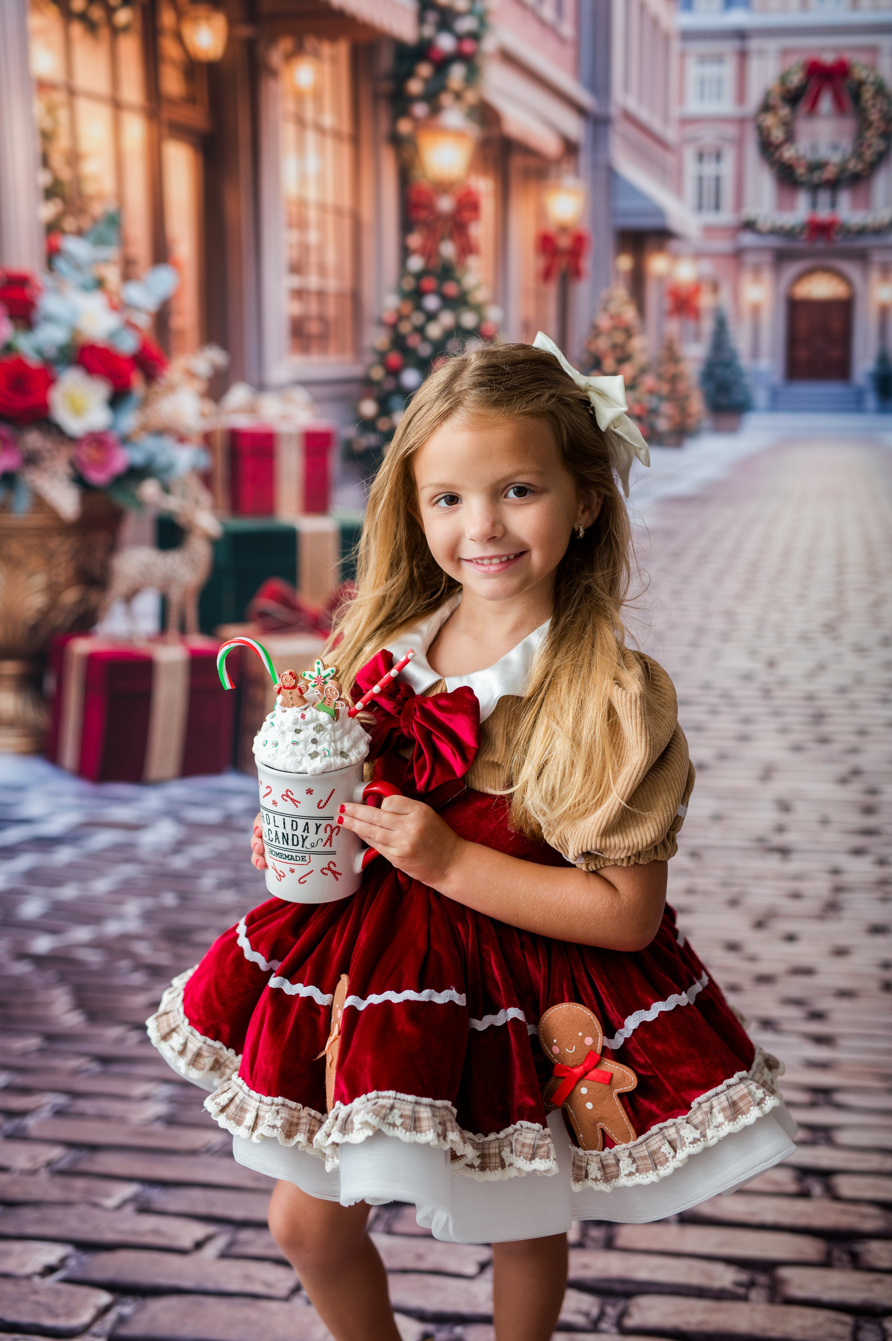 A young girl with long blonde hair wearing a festive red and white holiday dress, holding a 'Mistletoe Hot Chocolate' cup. The background is a blurred Christmas-themed street scene with lights, wreaths, and gifts.