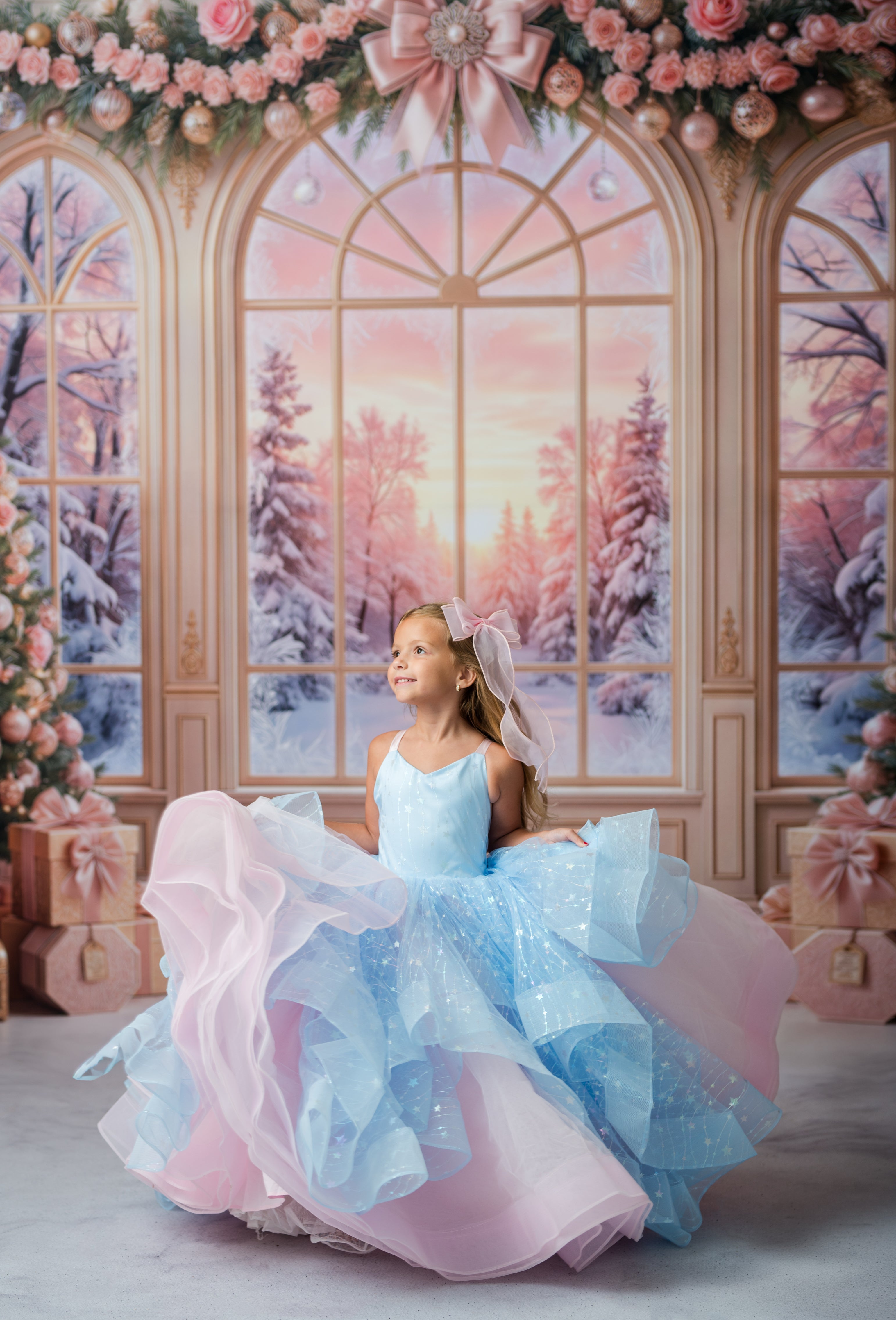 A young girl wearing a voluminous light blue and pink ball gown with sparkly details, posing in front of a festive holiday studio backdrop featuring arched windows with snowy winter scenery.