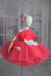 A pink tulle flower girl dress with a simple silhouette, featuring a white belt and bow, adjustable shoulder ties, and an elastic waist displayed on a mannequin.