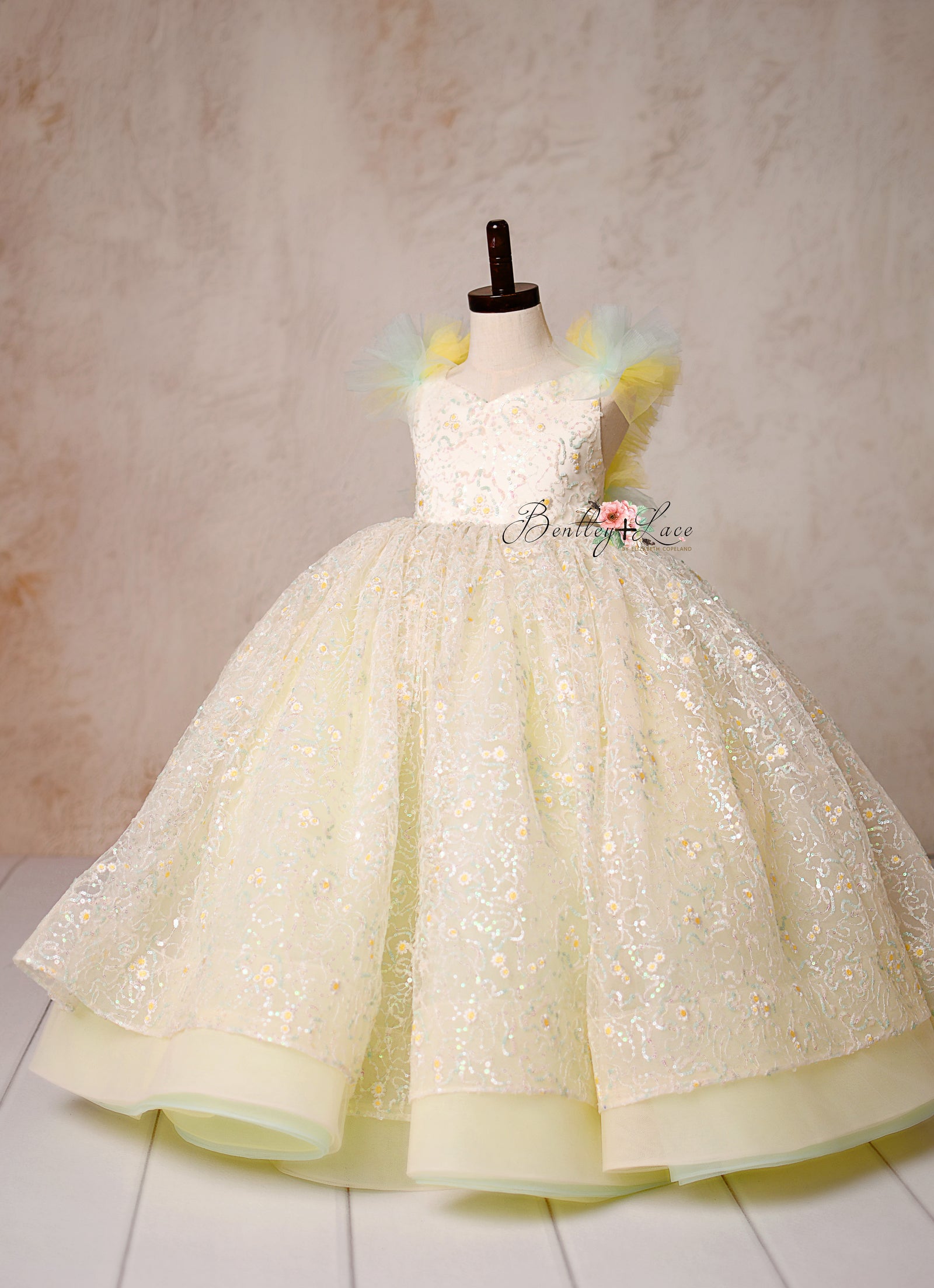 Capture timeless elegance with the Harmony Gown, featuring shimmering daisy lace, pastel hues, and a fairytale-inspired silhouette.

