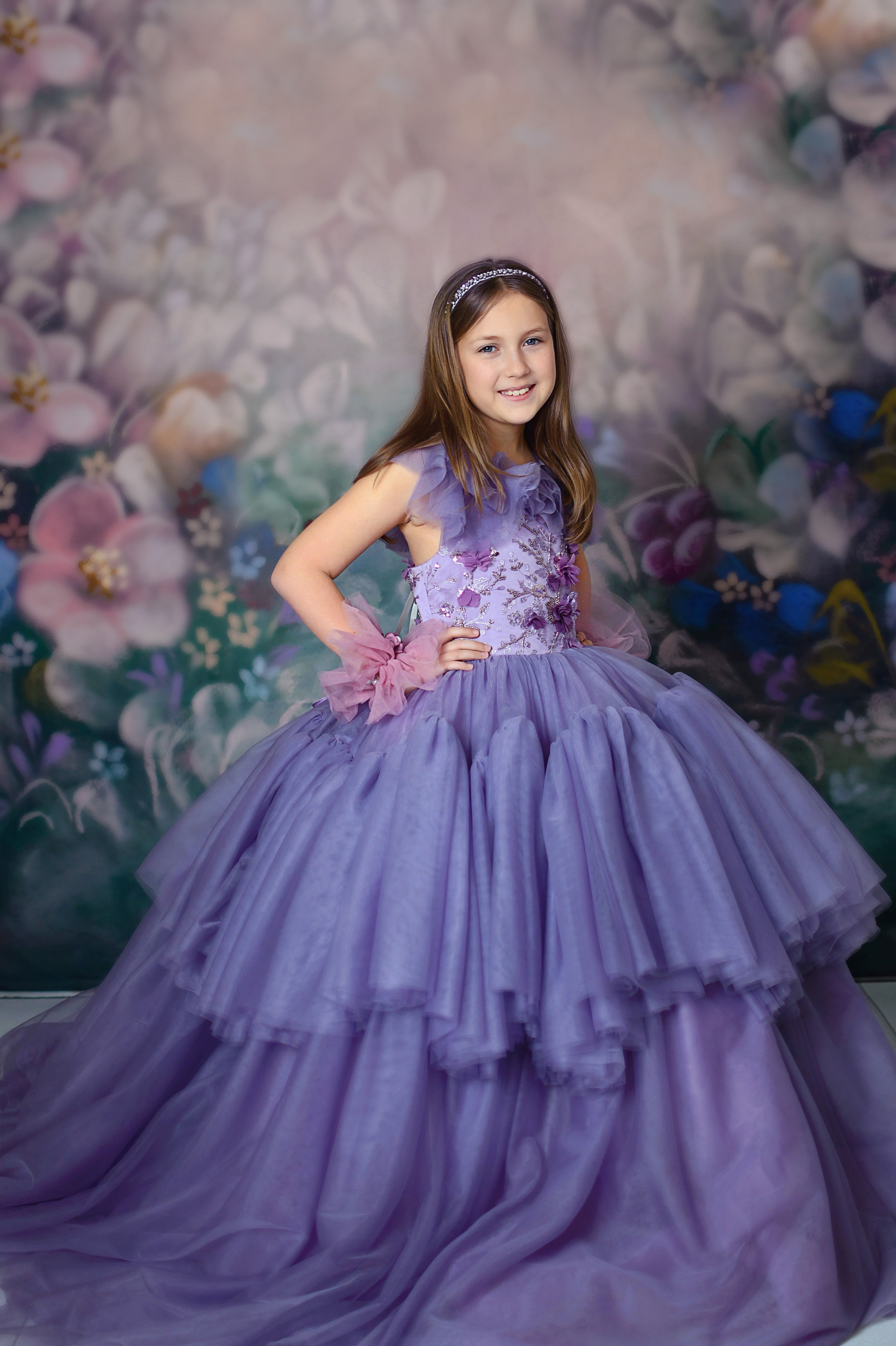 "Girl in a violet floor-length gown with floral appliqués and layered tulle skirt, posing elegantly against a floral backdrop."
