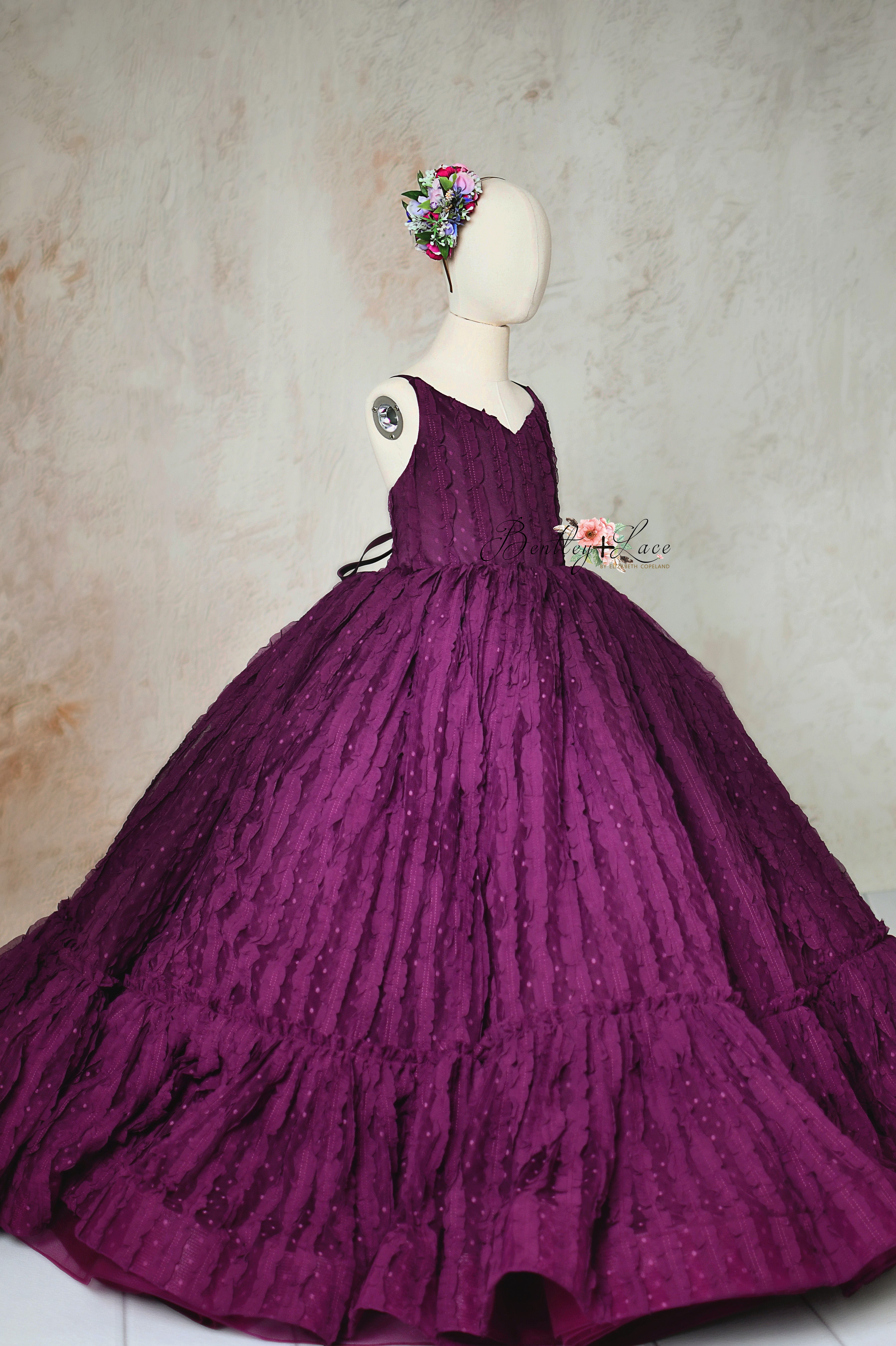 A luxurious deep plum ball gown displayed on a child-sized mannequin, featuring textured lace fabric and a voluminous skirt.
