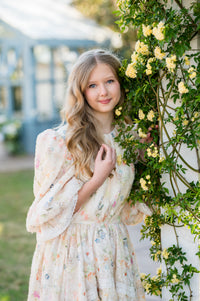 Young woman in a floral dress standing next to a wall with yellow flowers outdoors.