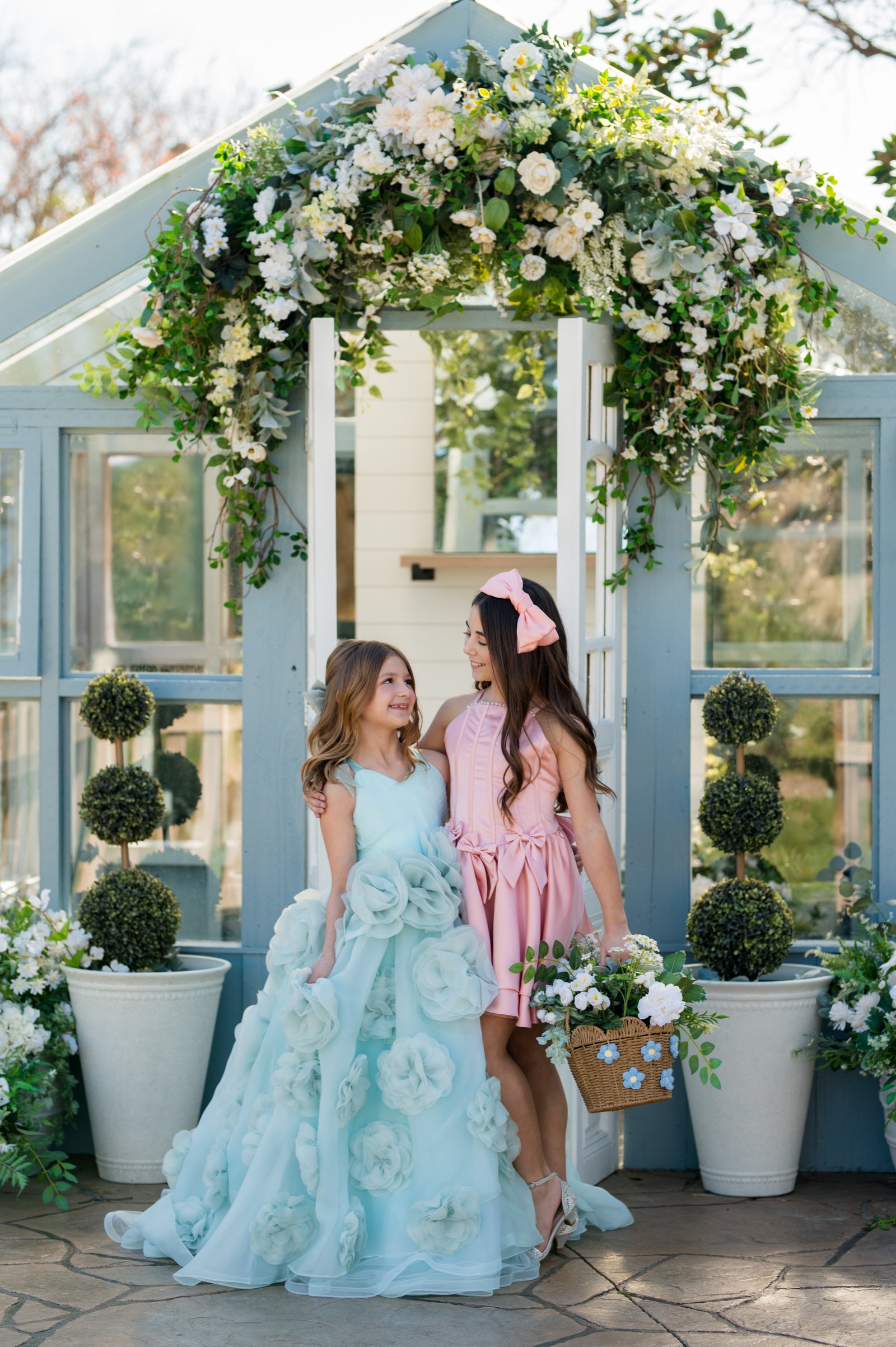 Two women in formal dresses standing under a floral archway with decorative plants around.