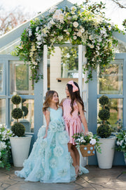 Two women in formal dresses standing under a floral archway with decorative plants around.