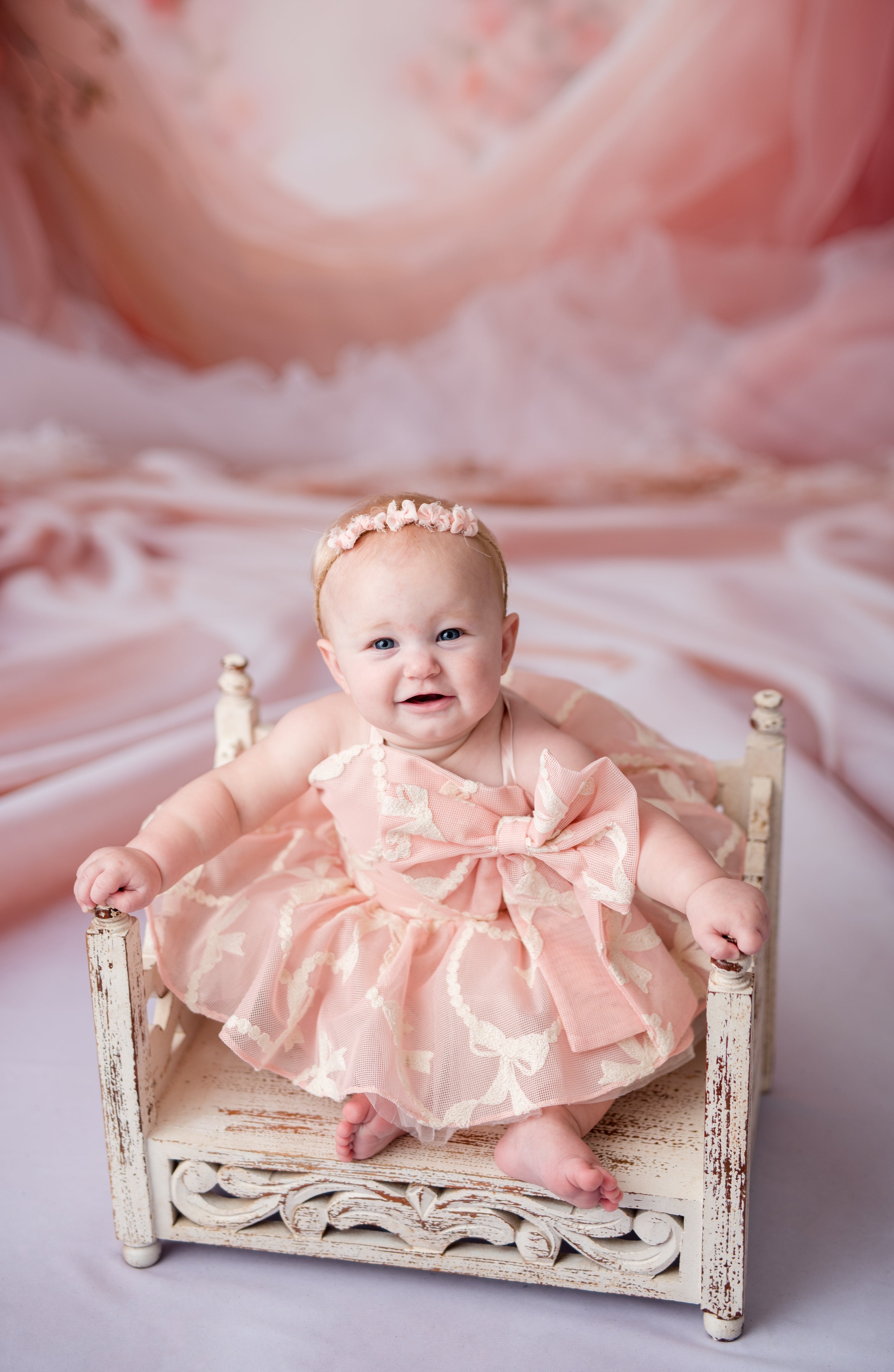 Baby in a pink dress sitting on a small bed with a soft pink background