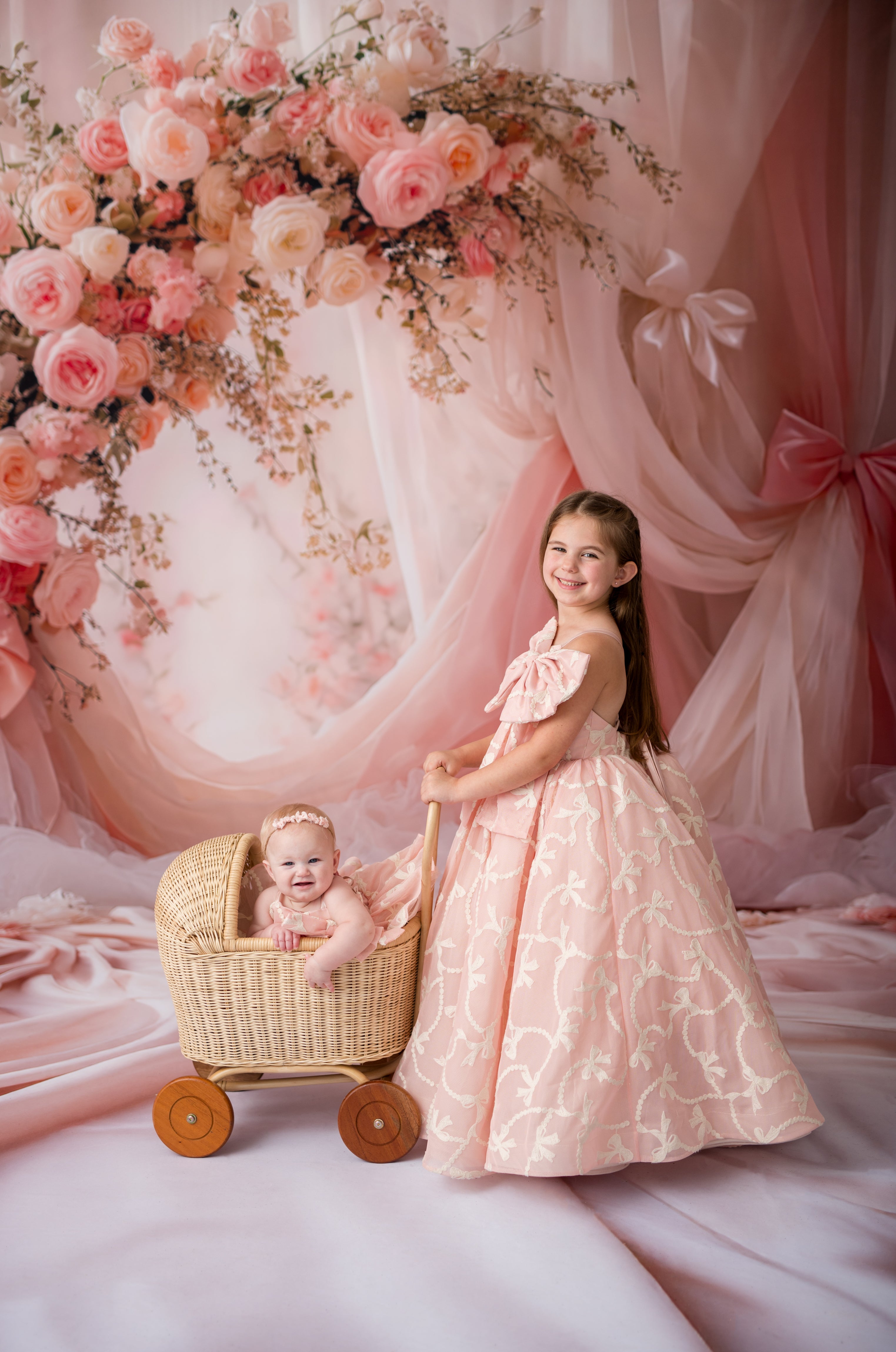 Two young girls in pink dresses with a baby in a stroller against a floral and pink curtain backdrop.