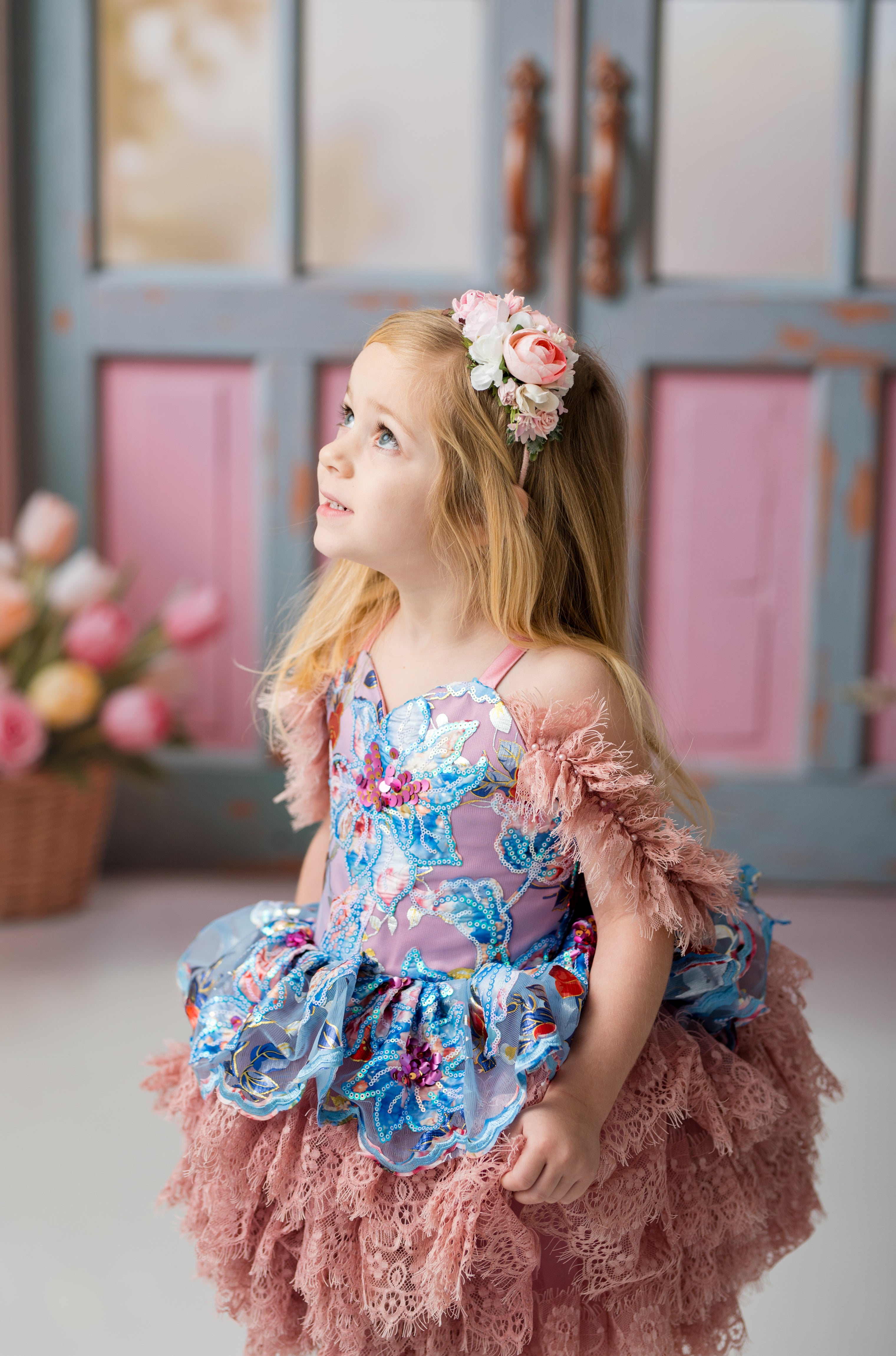 Young girl in a floral dress with a decorative headband, standing in a softly blurred indoor setting.