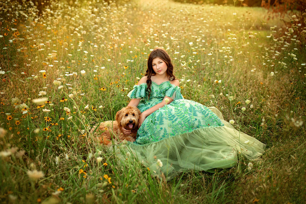Young girl in a green dress sitting in a field with a dog, surrounded by tall grass and wildflowers.
