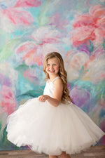 A young girl wearing a white tulle dress with a satin bodice, standing in front of a colorful floral backdrop.