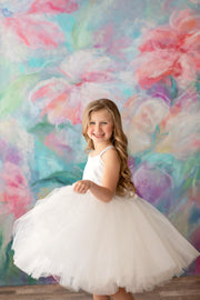 A young girl wearing a white tulle dress with a satin bodice, standing in front of a colorful floral backdrop.