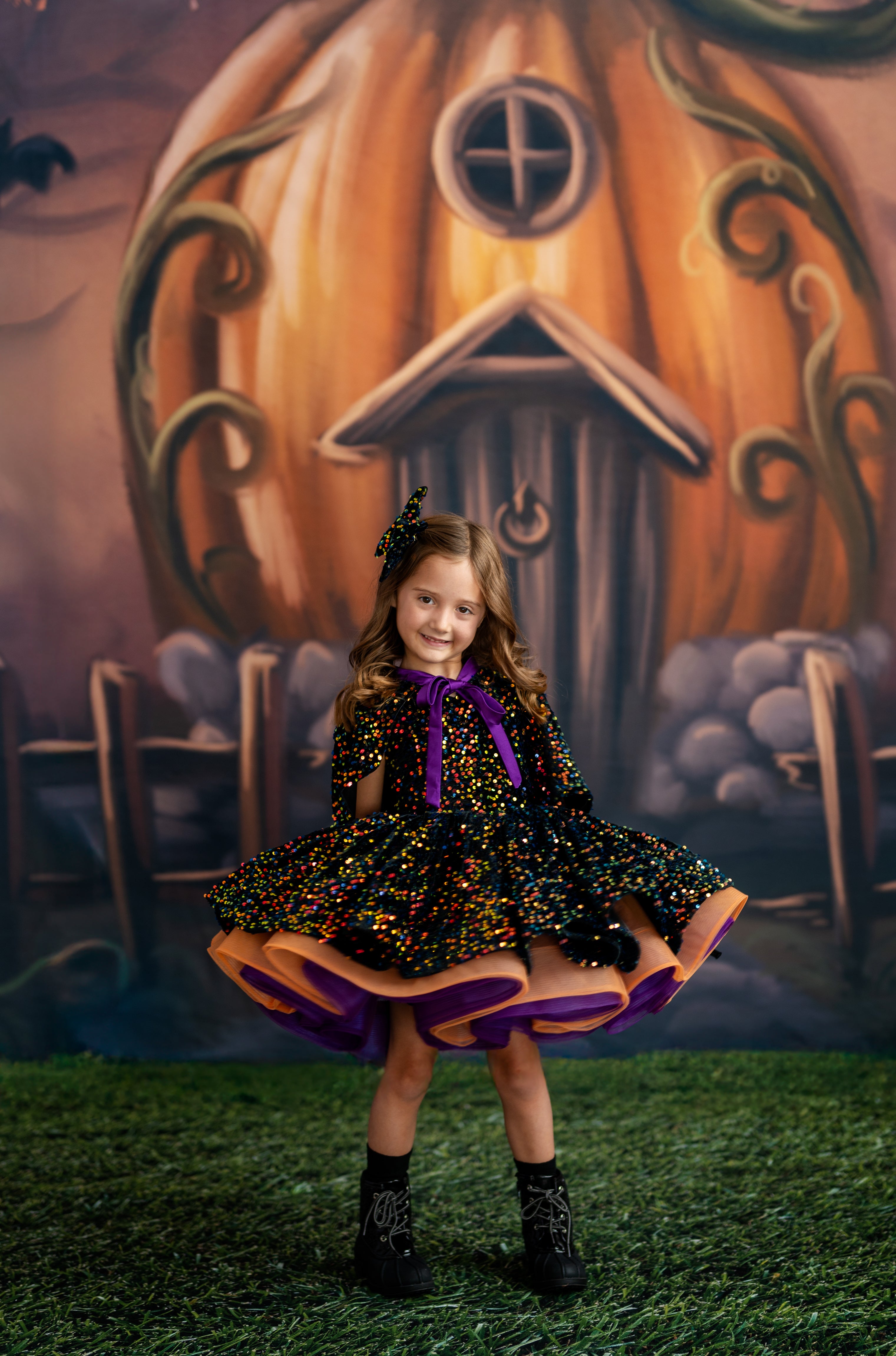 Young girl in a Halloween-themed dress standing in front of a pumpkin house backdrop.