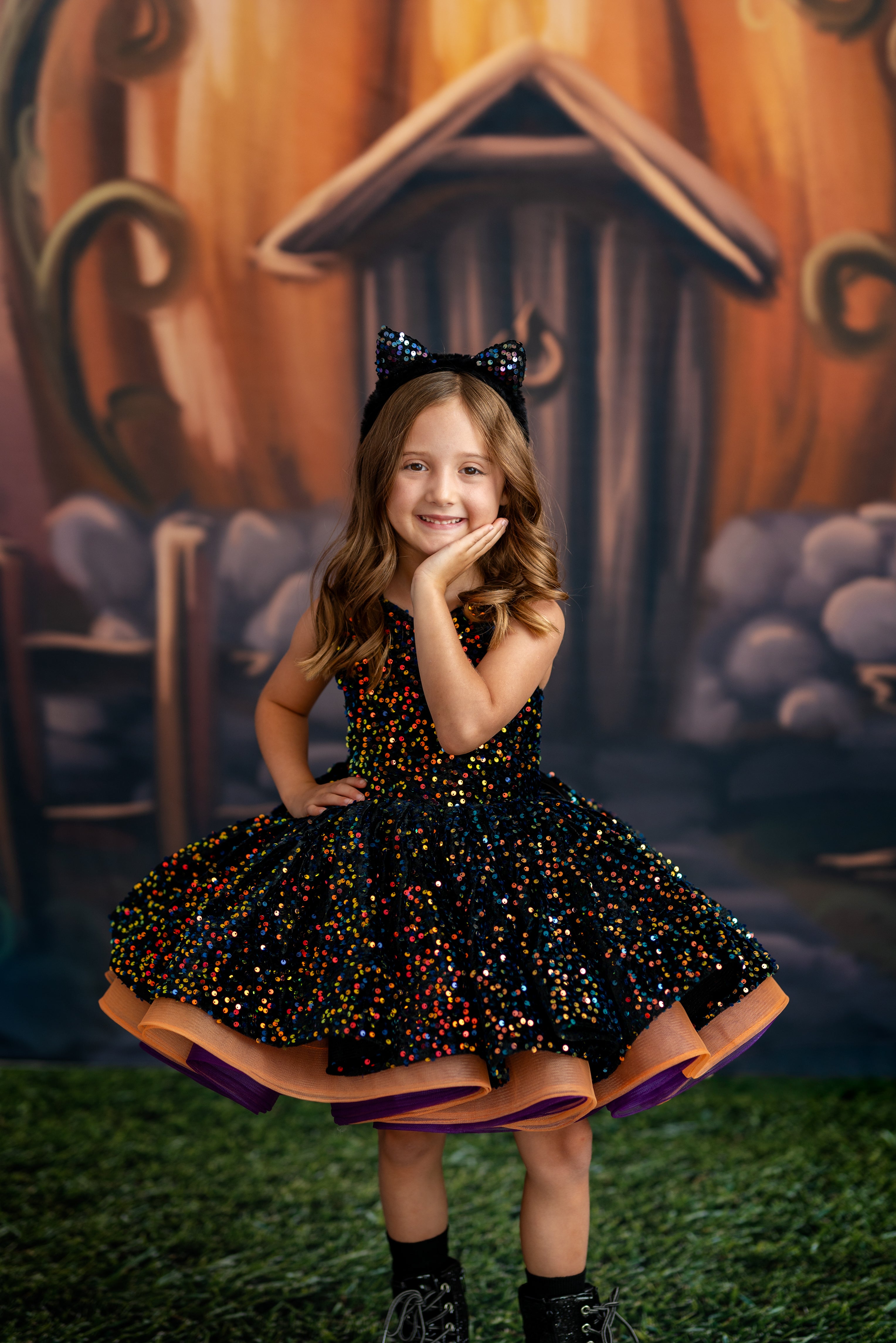 Young girl in a Halloween-themed dress posing in front of a pumpkin house backdrop.