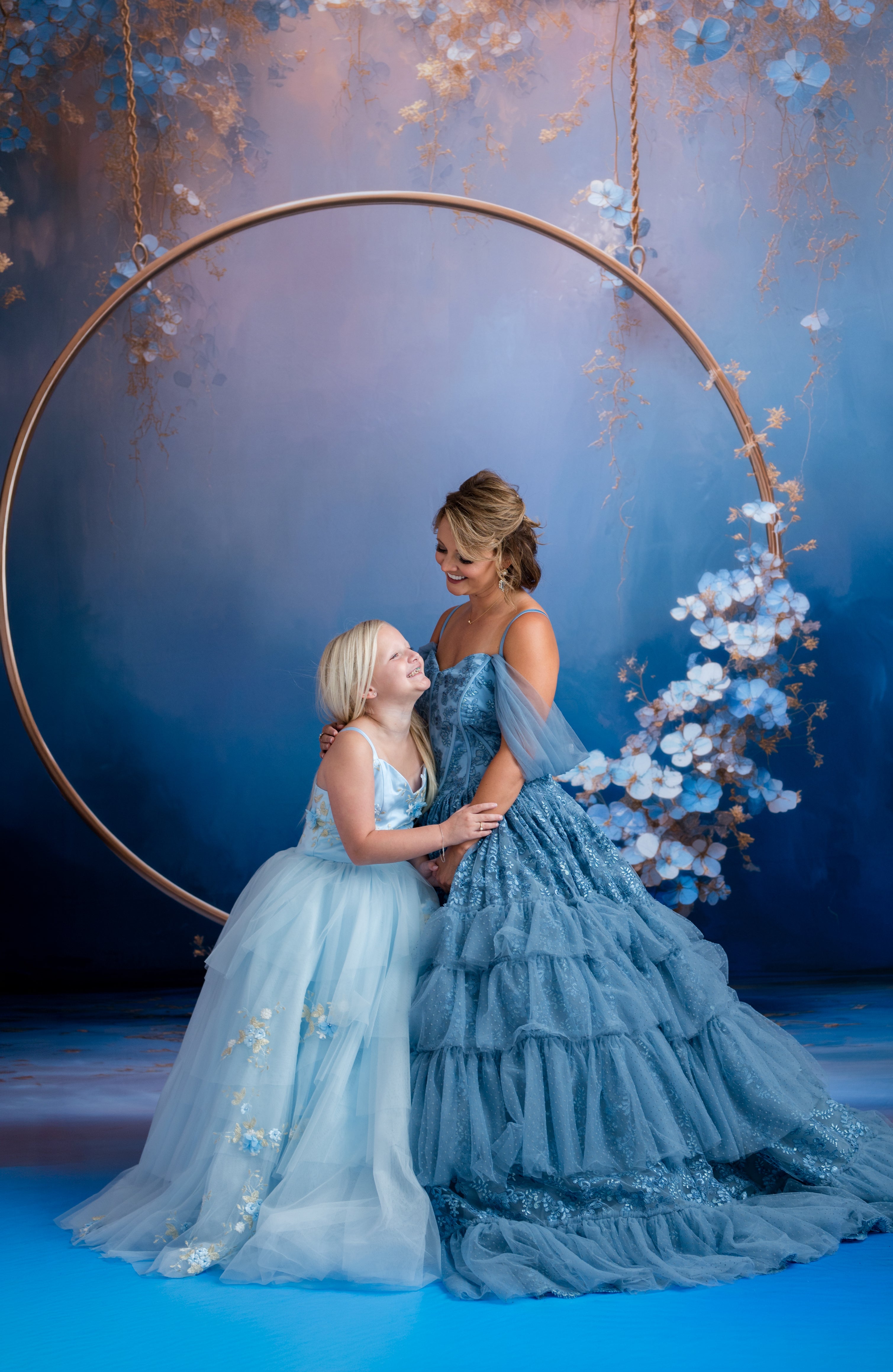 Two women in elegant blue dresses standing under a large circular frame with floral decorations against a blue background.