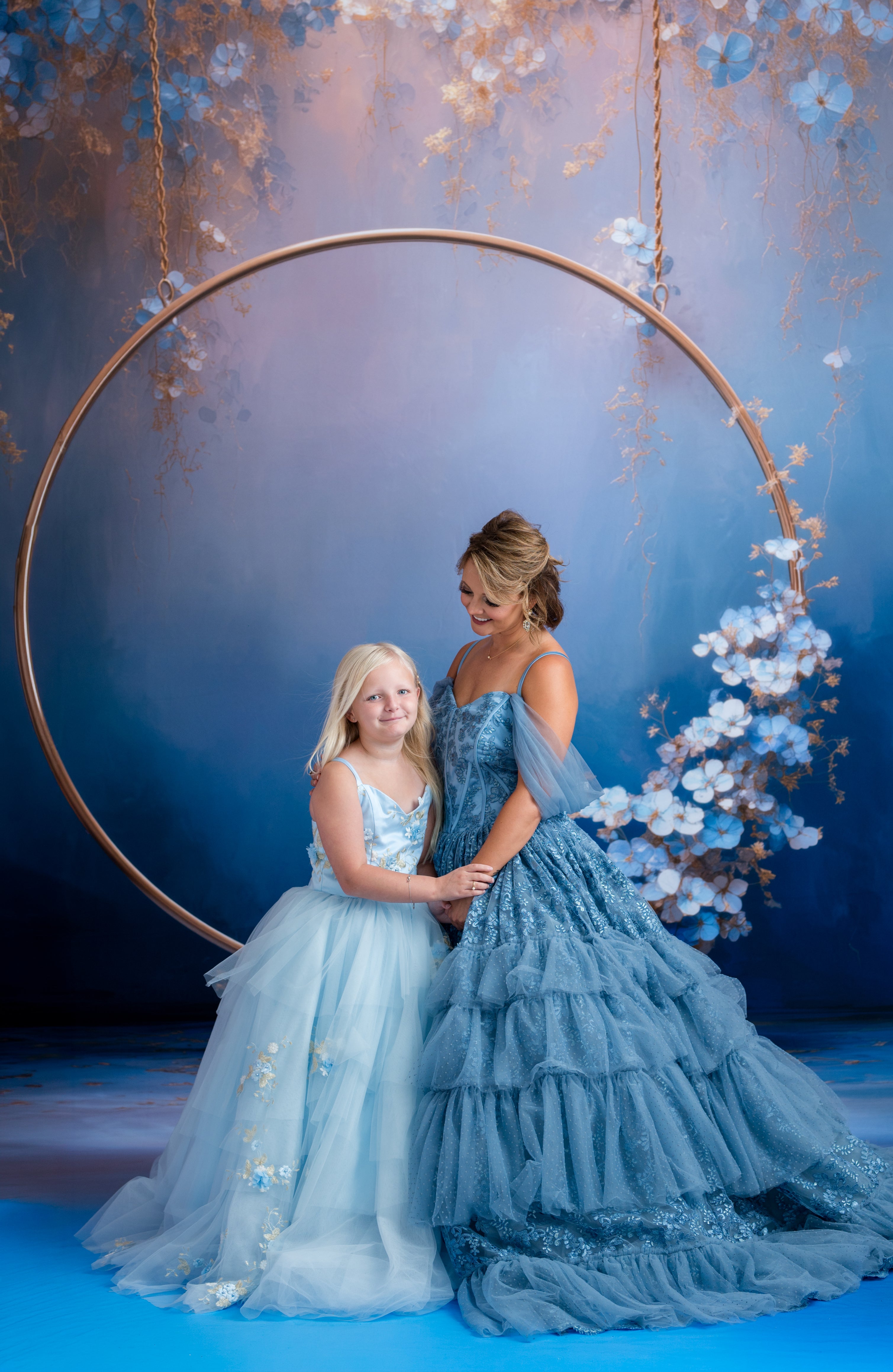 Two women in elegant blue dresses standing under a floral arch with a blue background.