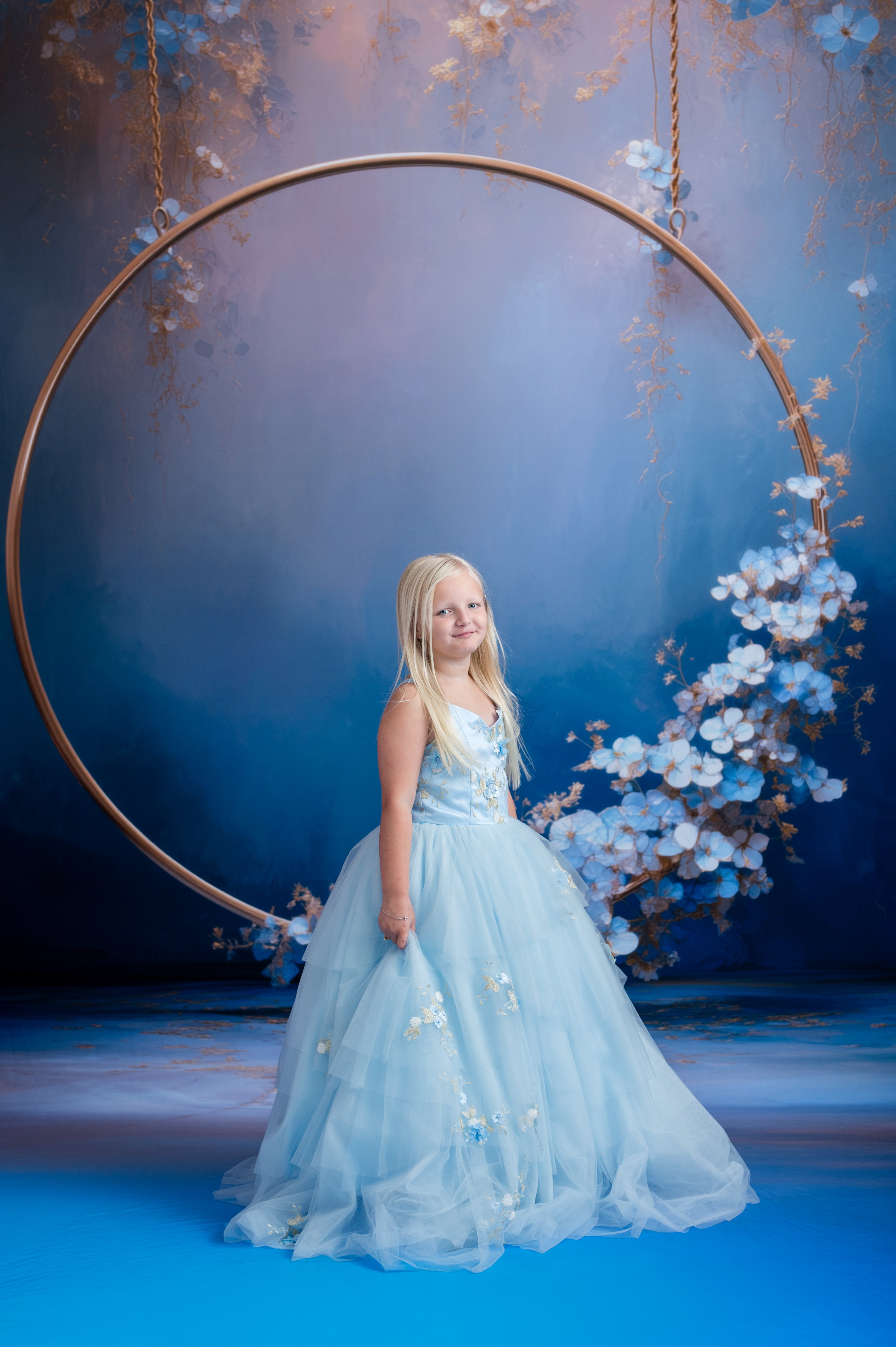Young girl in a light blue dress standing in front of a large circular frame with floral decorations against a blue background.