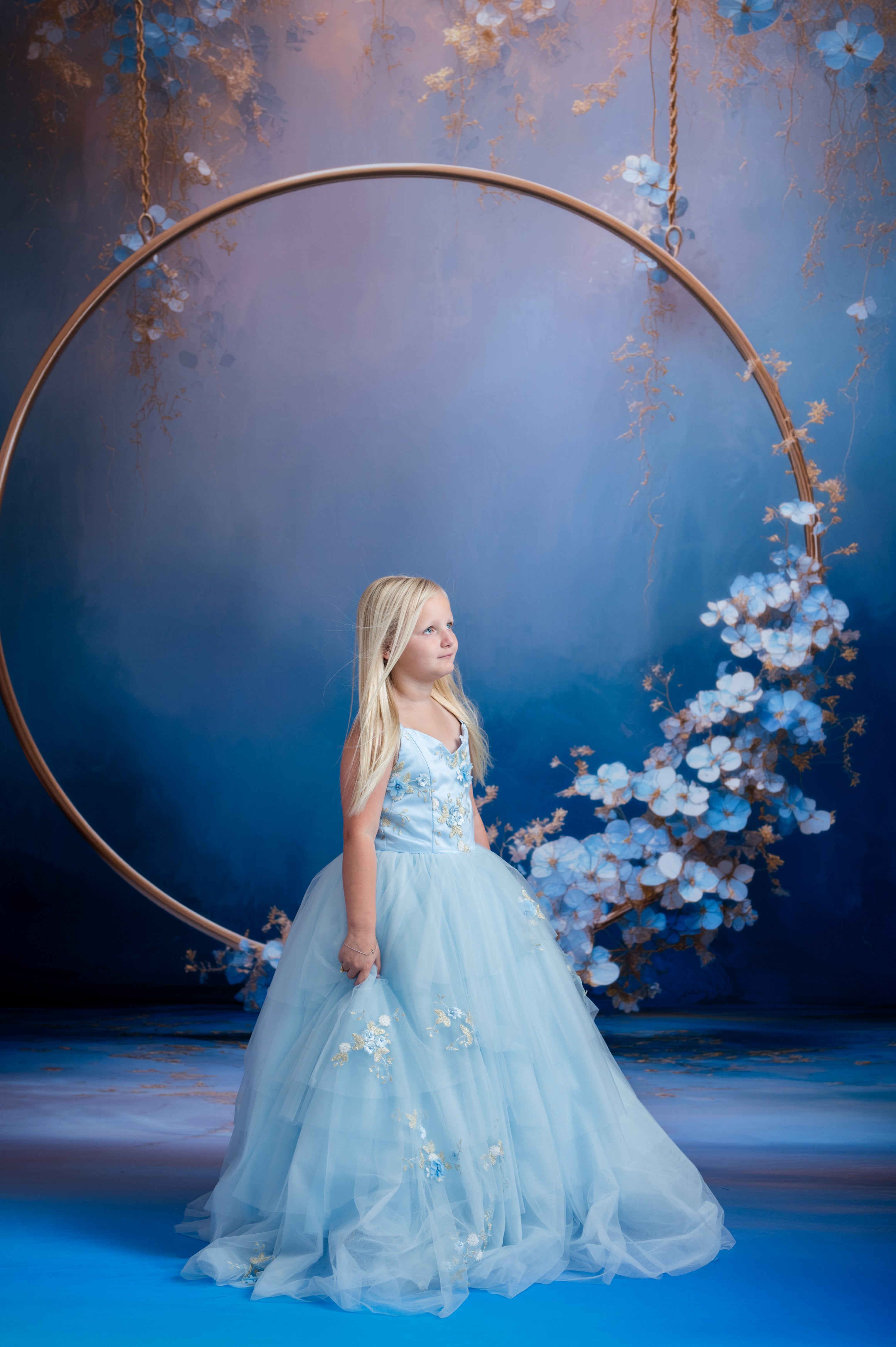 Young girl in a light blue dress standing under a floral arch with a blue background
