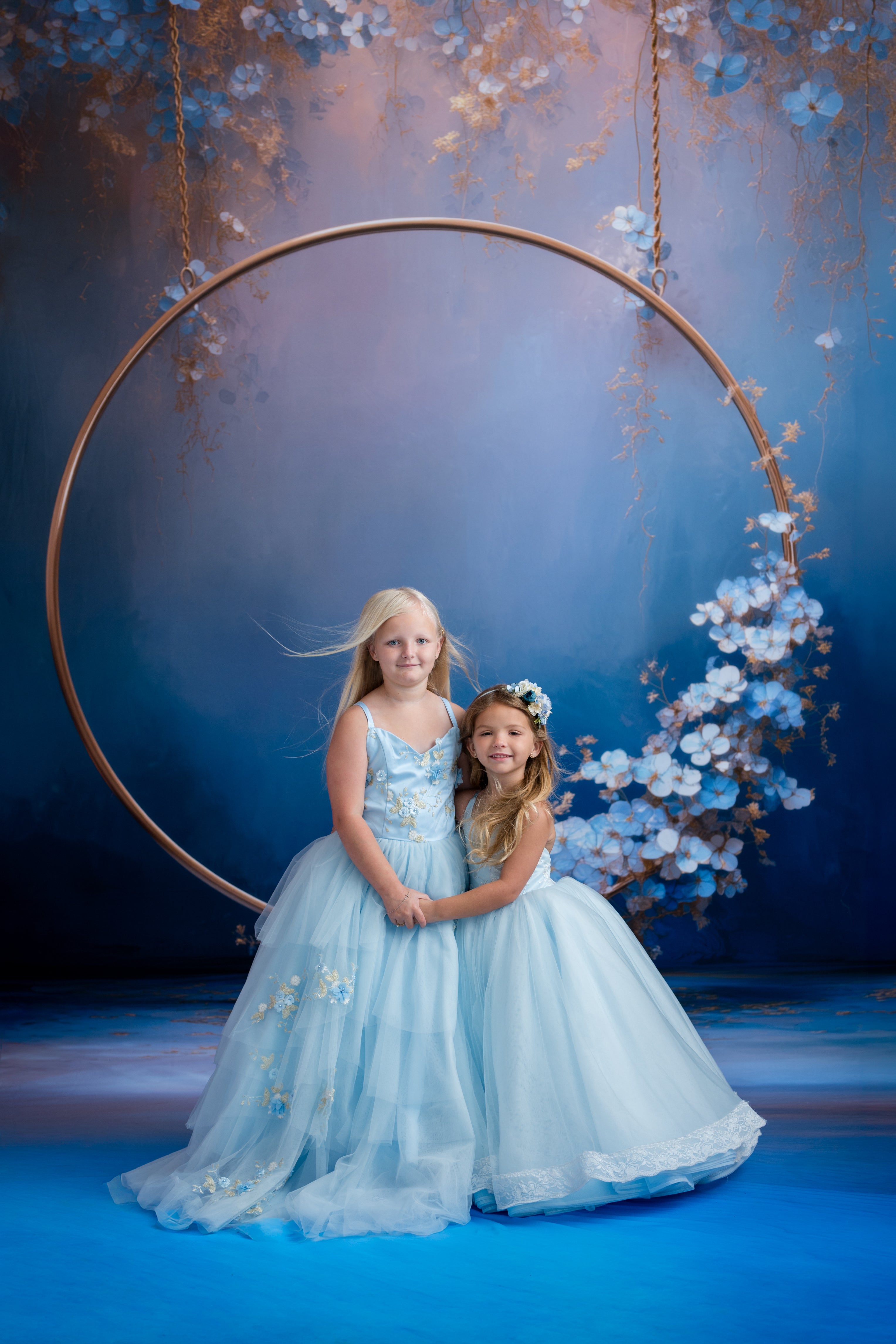 Two young girls in light blue dresses standing under a floral arch with a blue background.