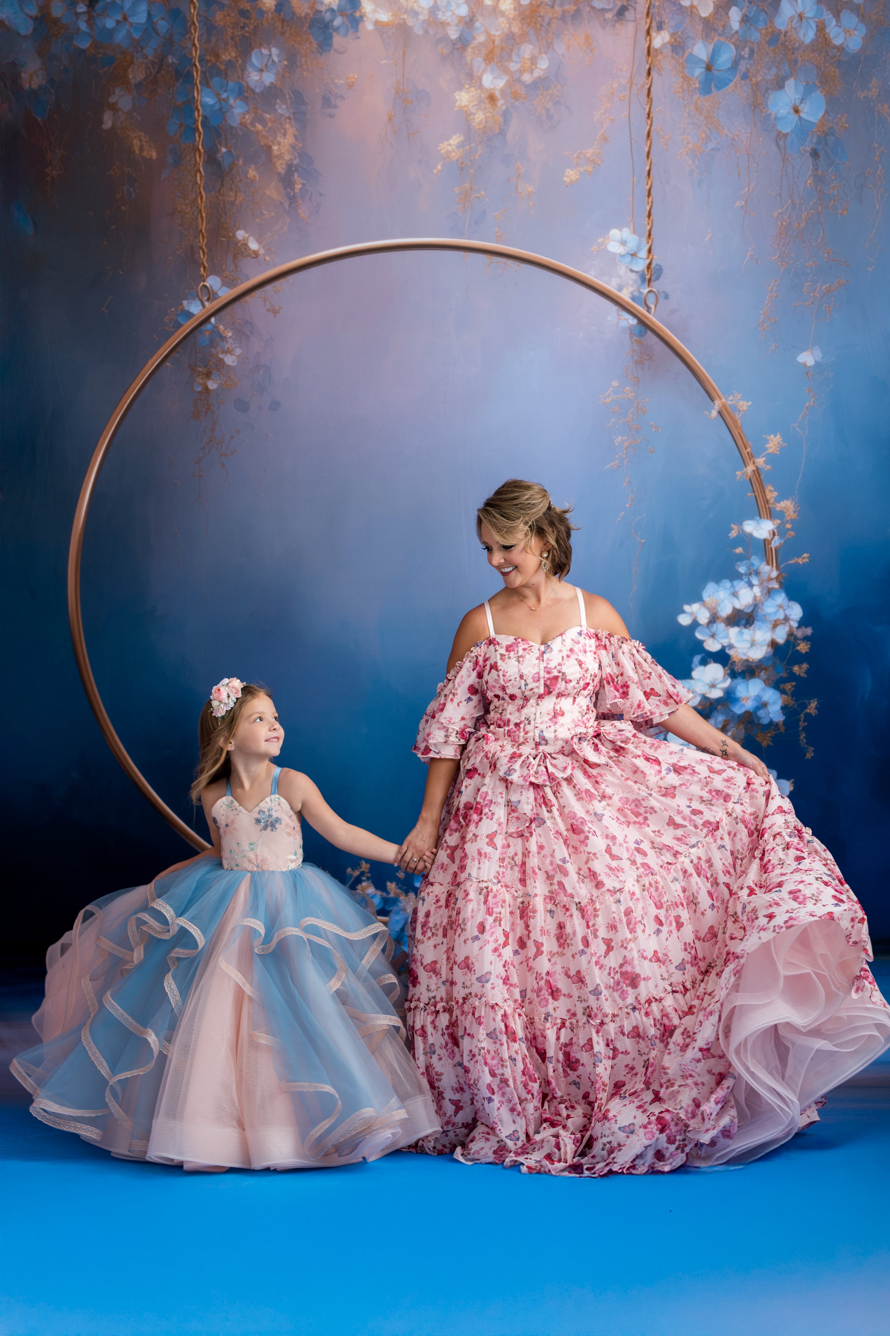 Two women in floral dresses standing under a circular metal frame against a blue background with floral decorations.