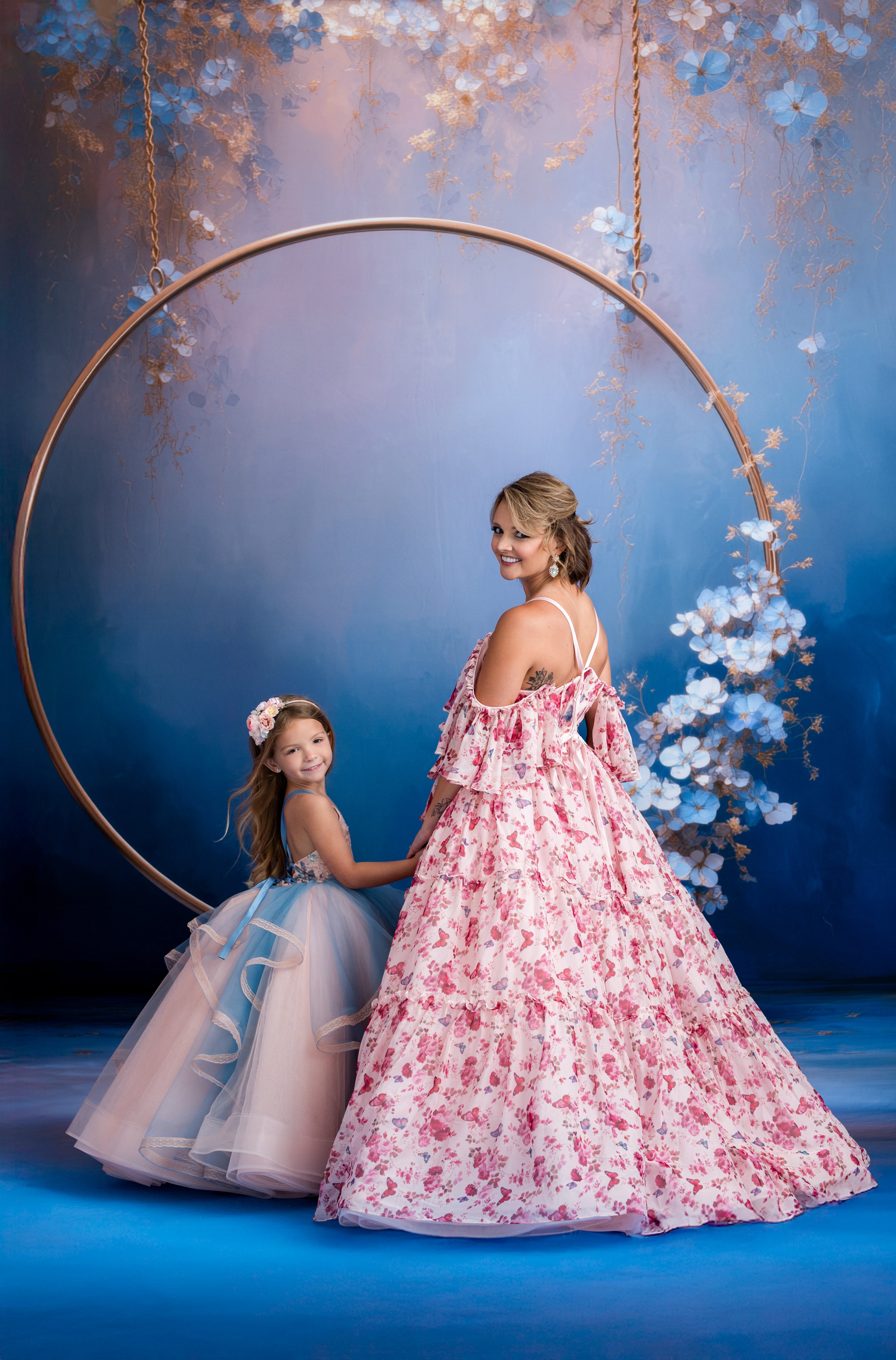 Two women in elegant dresses standing behind a floral hoop against a blue background