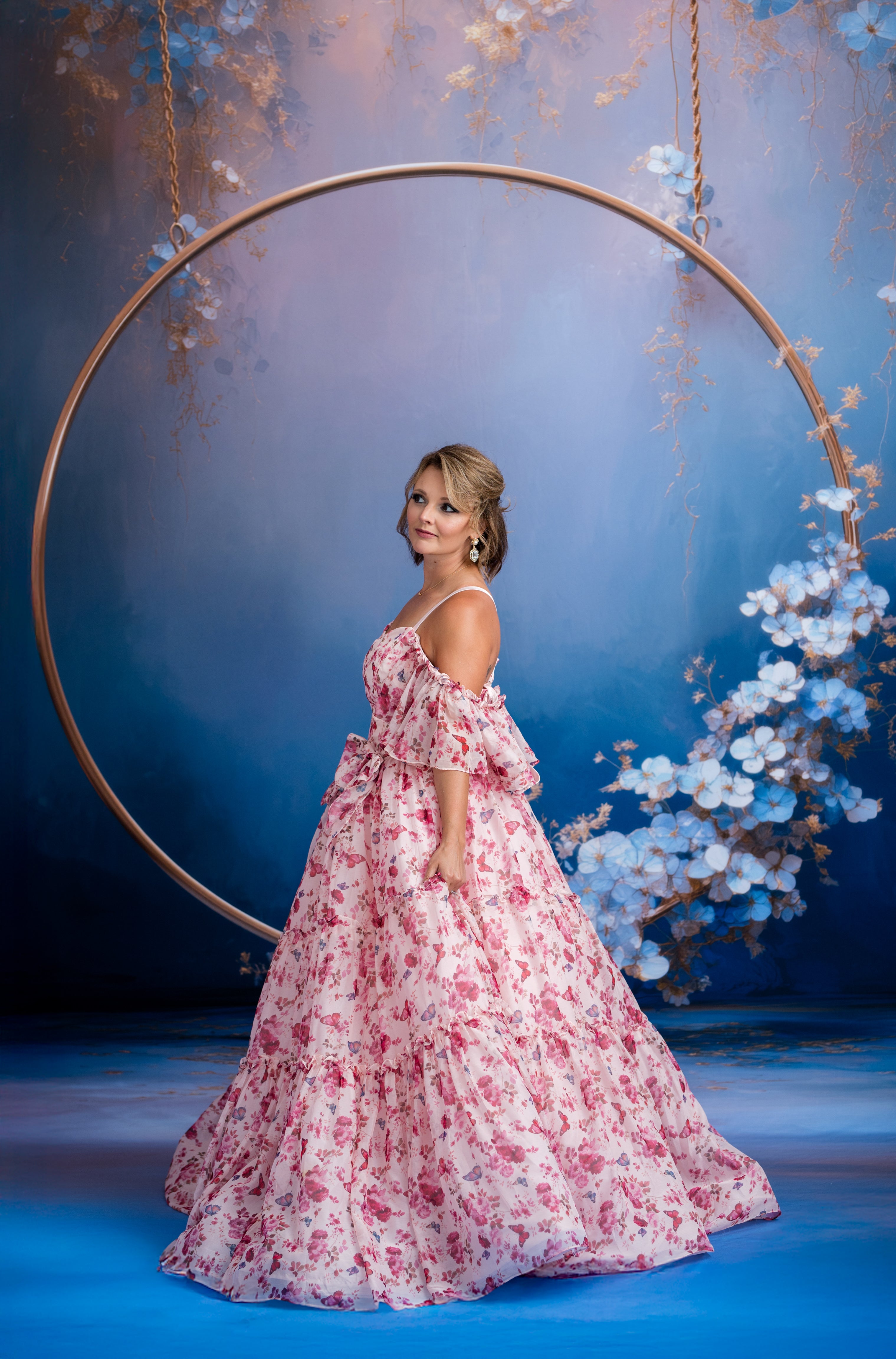 Woman in a floral dress standing in front of a decorative circular frame with flowers against a blue background