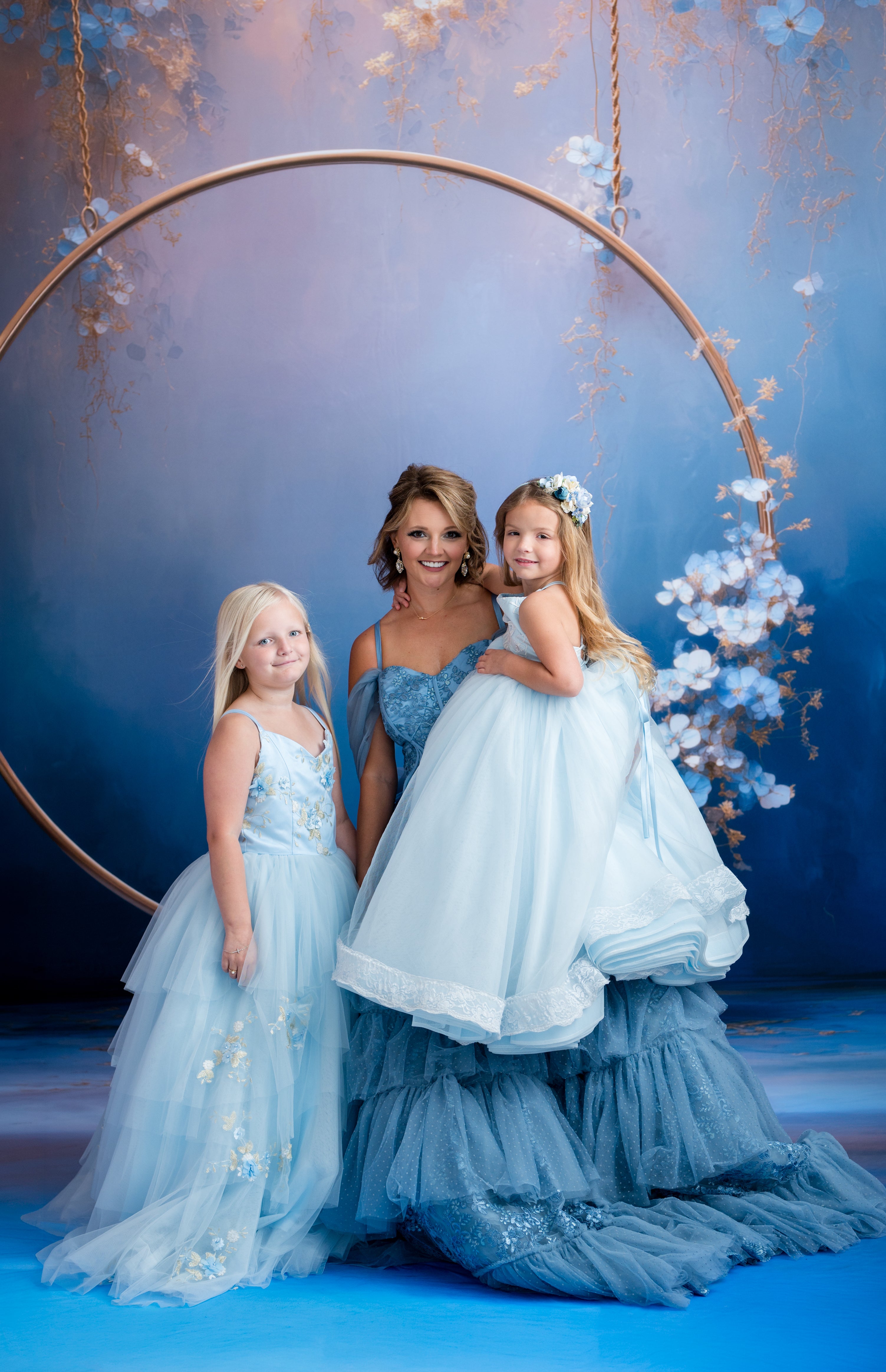 Woman and two young girls in matching blue dresses standing in front of a floral hoop against a blue background