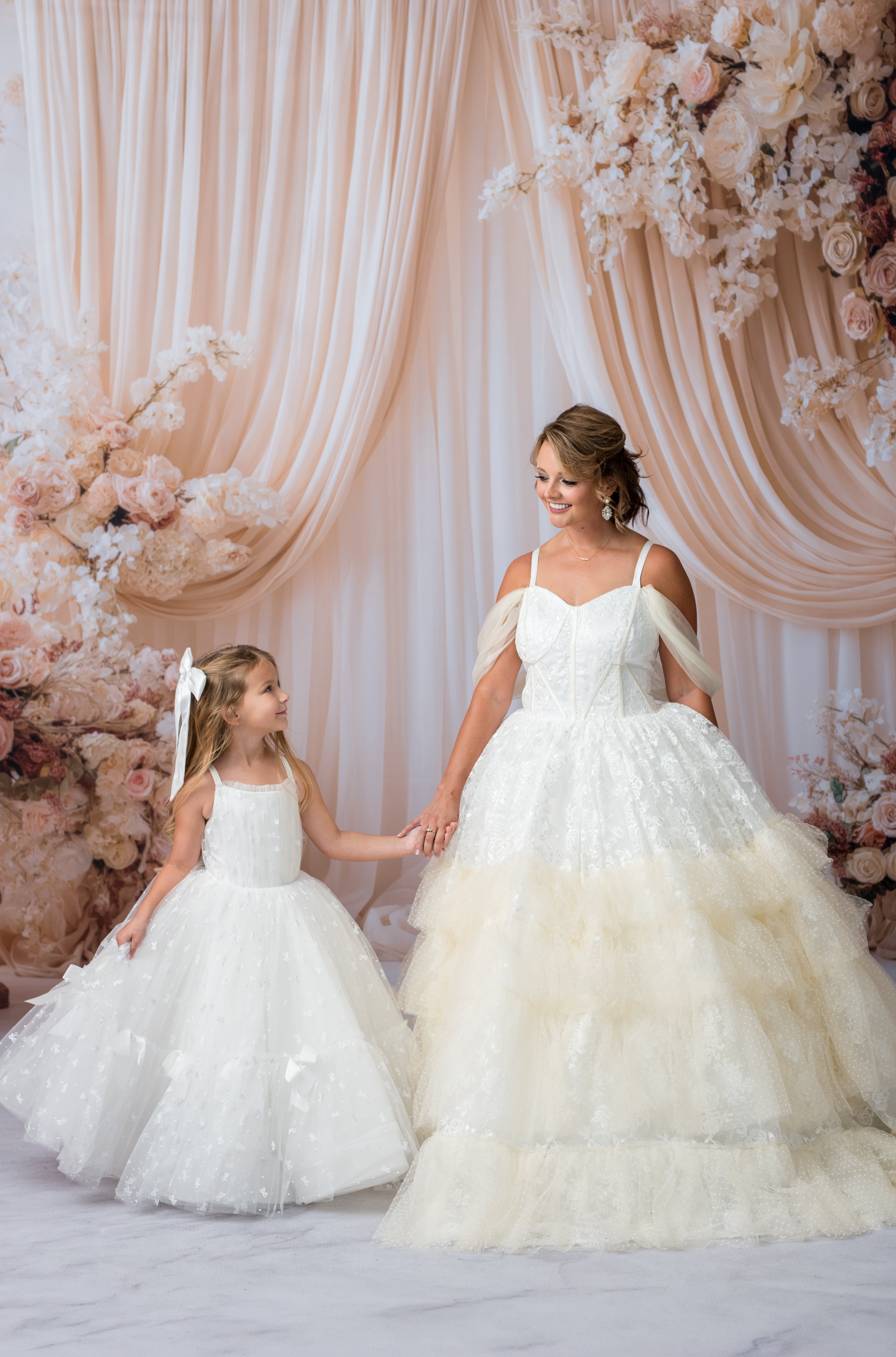 Two women in white dresses standing in front of a floral and curtain backdrop