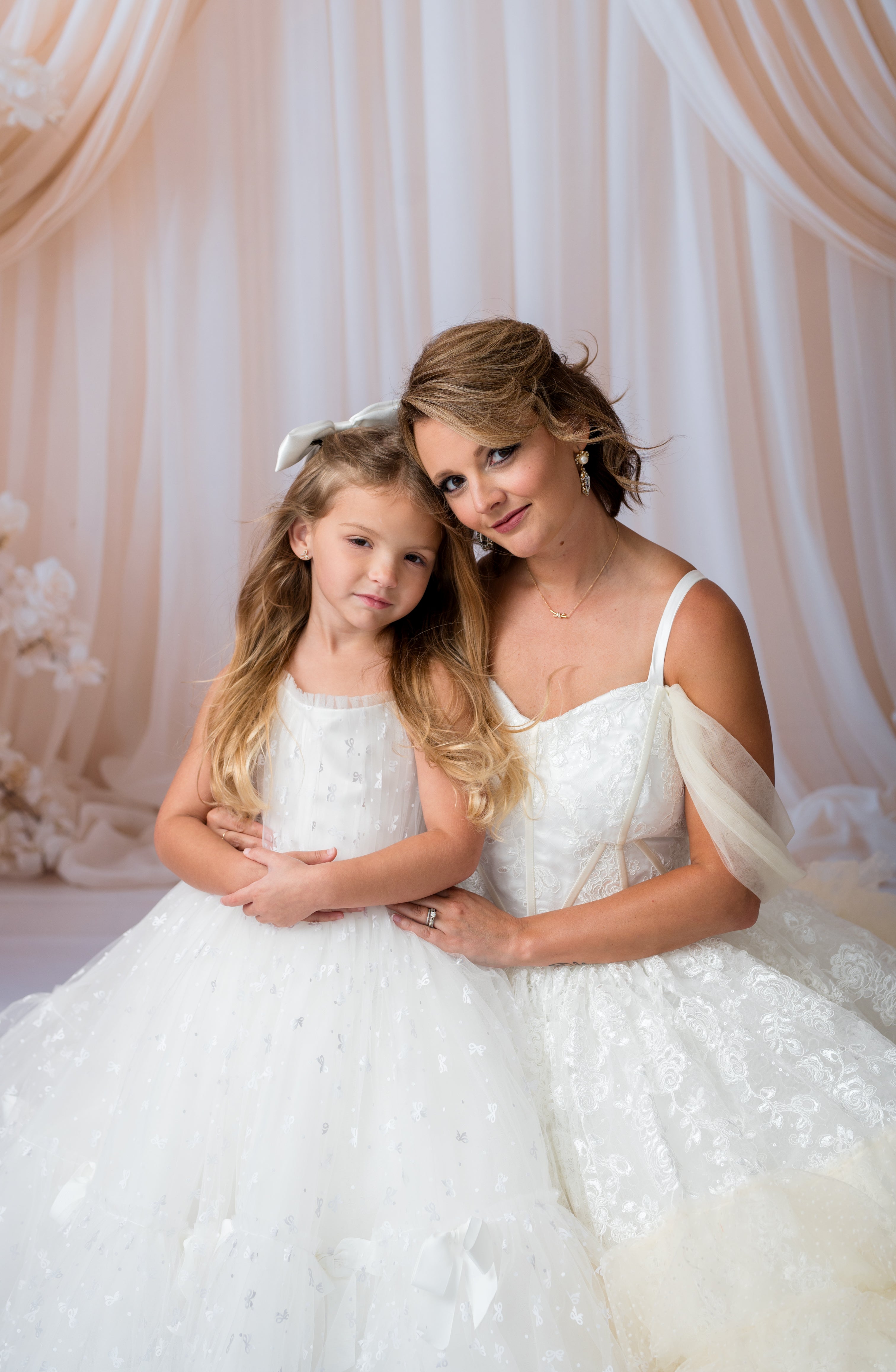 Woman in a white dress holding a young girl in a matching white dress against a soft pink curtain.