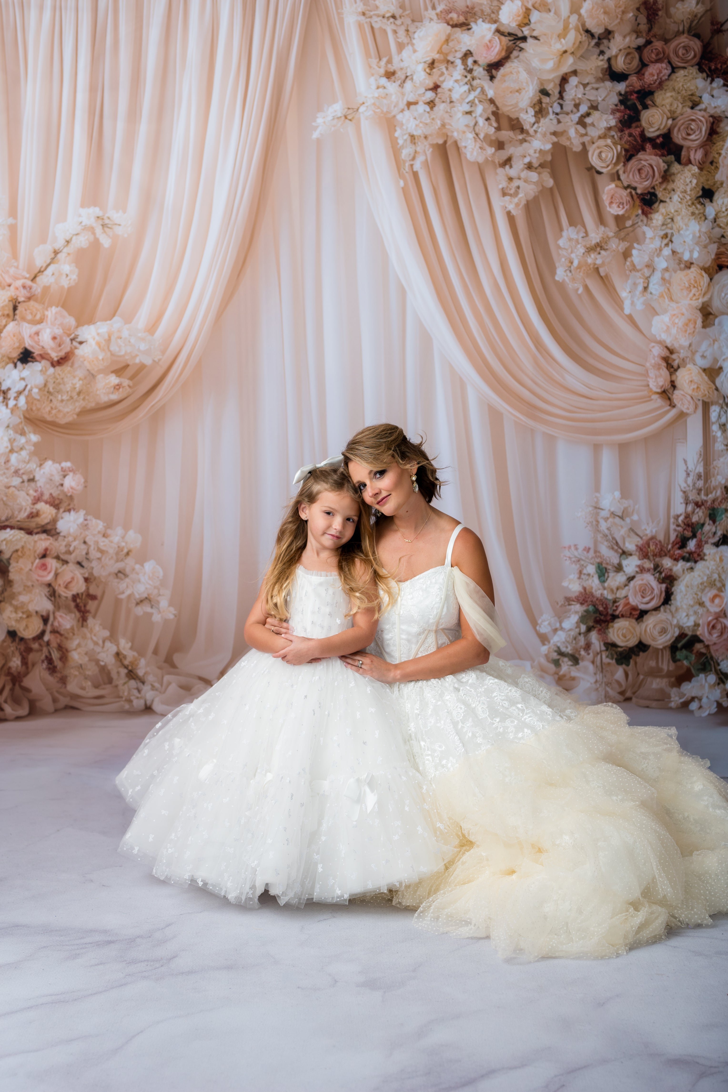Woman and young girl in white dresses standing in front of a floral and curtain backdrop