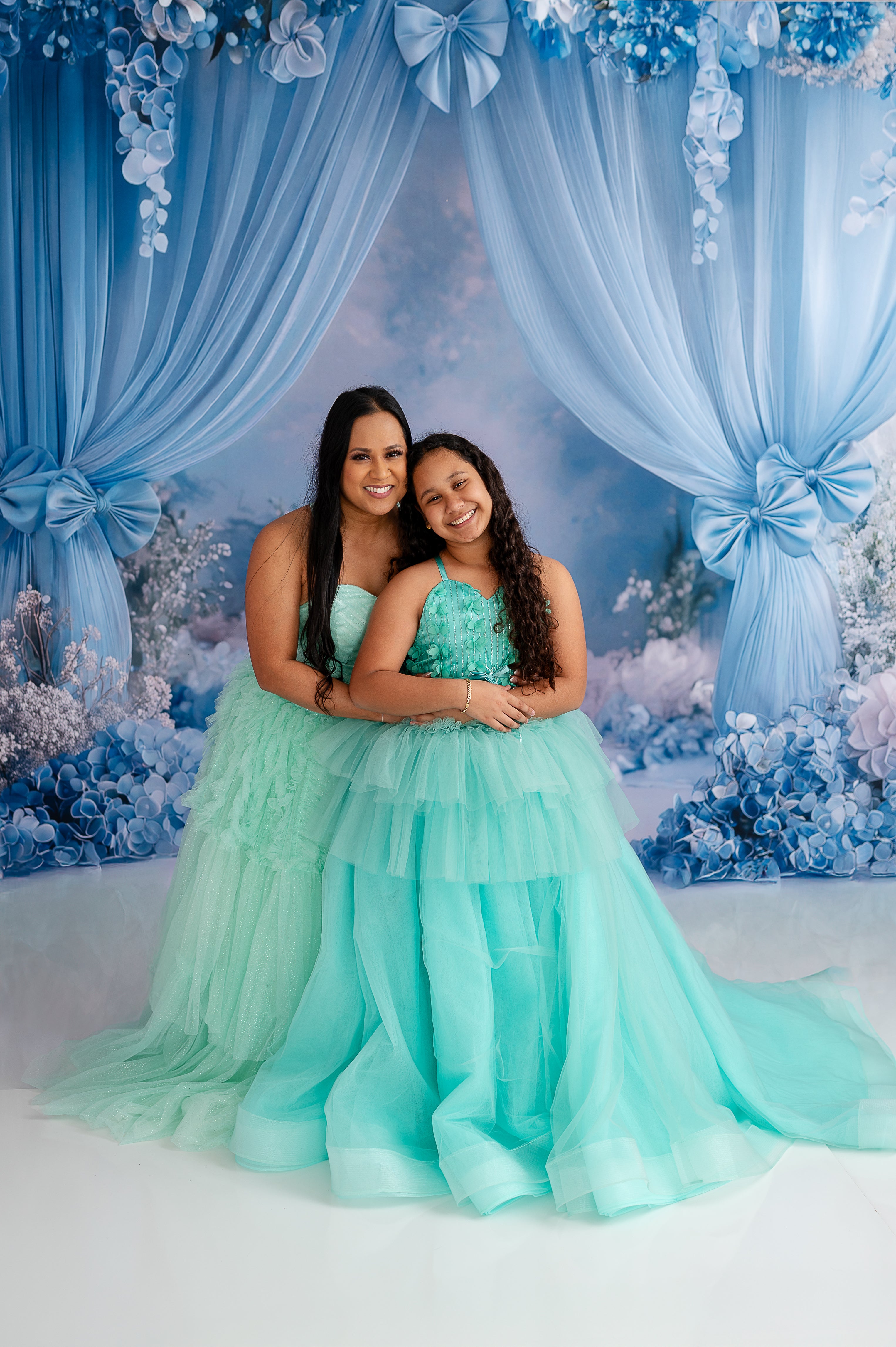 "Smiling mother and daughter wearing matching layered tulle gowns in shades of turquoise and mint."
