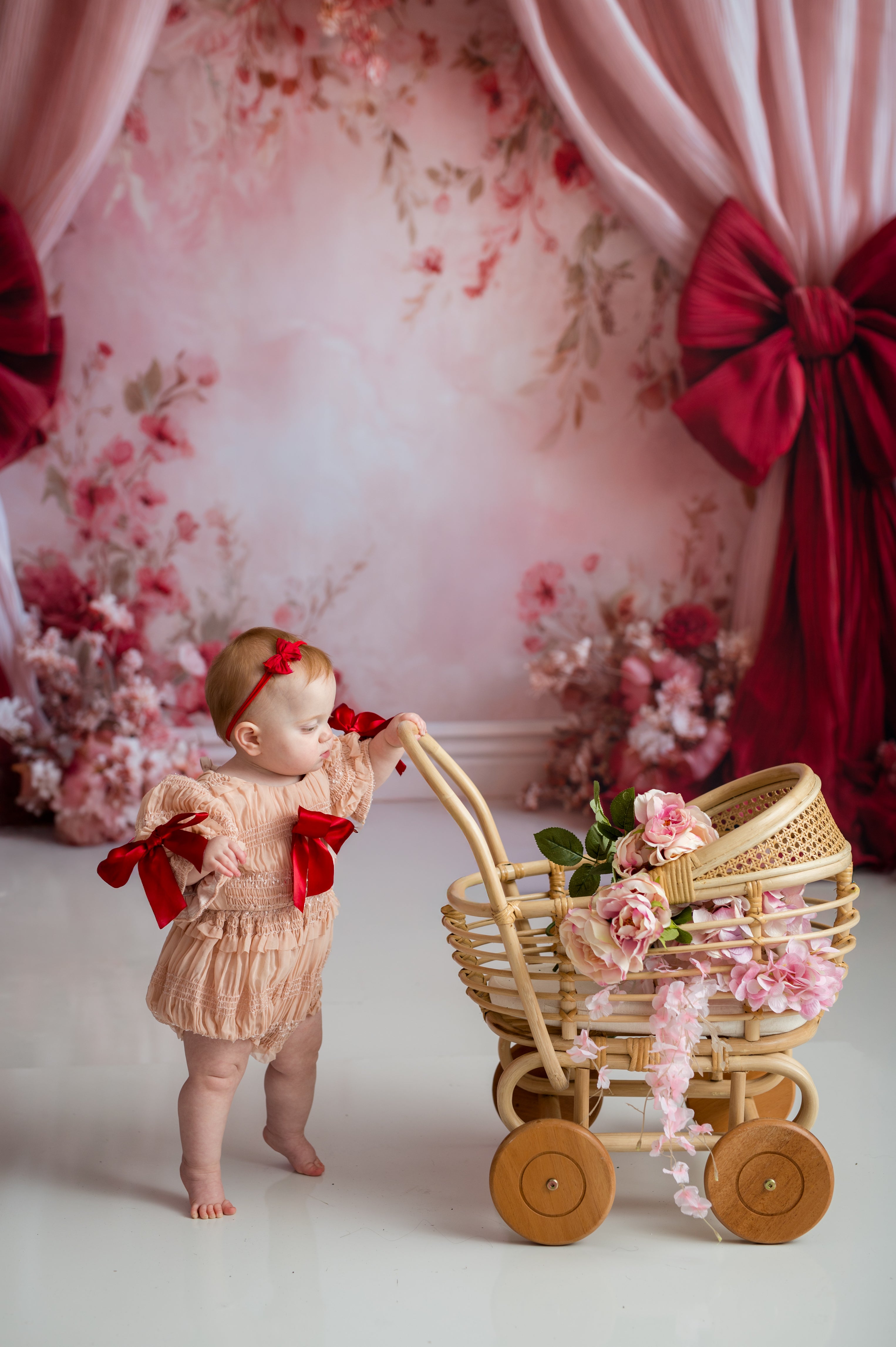 Baby in a beige outfit with red ribbons standing next to a decorative wicker cart against a floral backdrop.