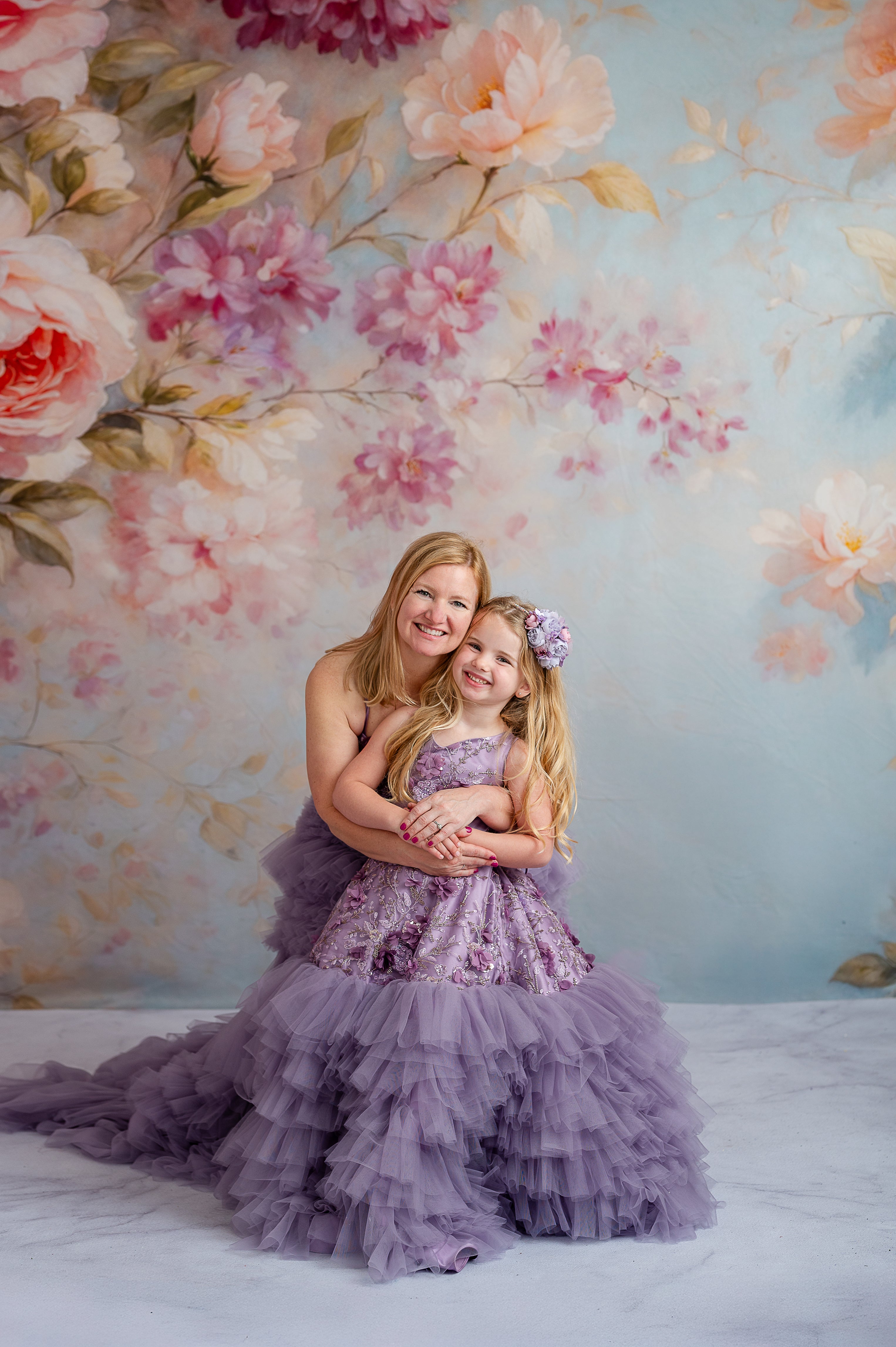 Mother holding hands with daughter wearing floor-length lavender floral tulle dresses.

