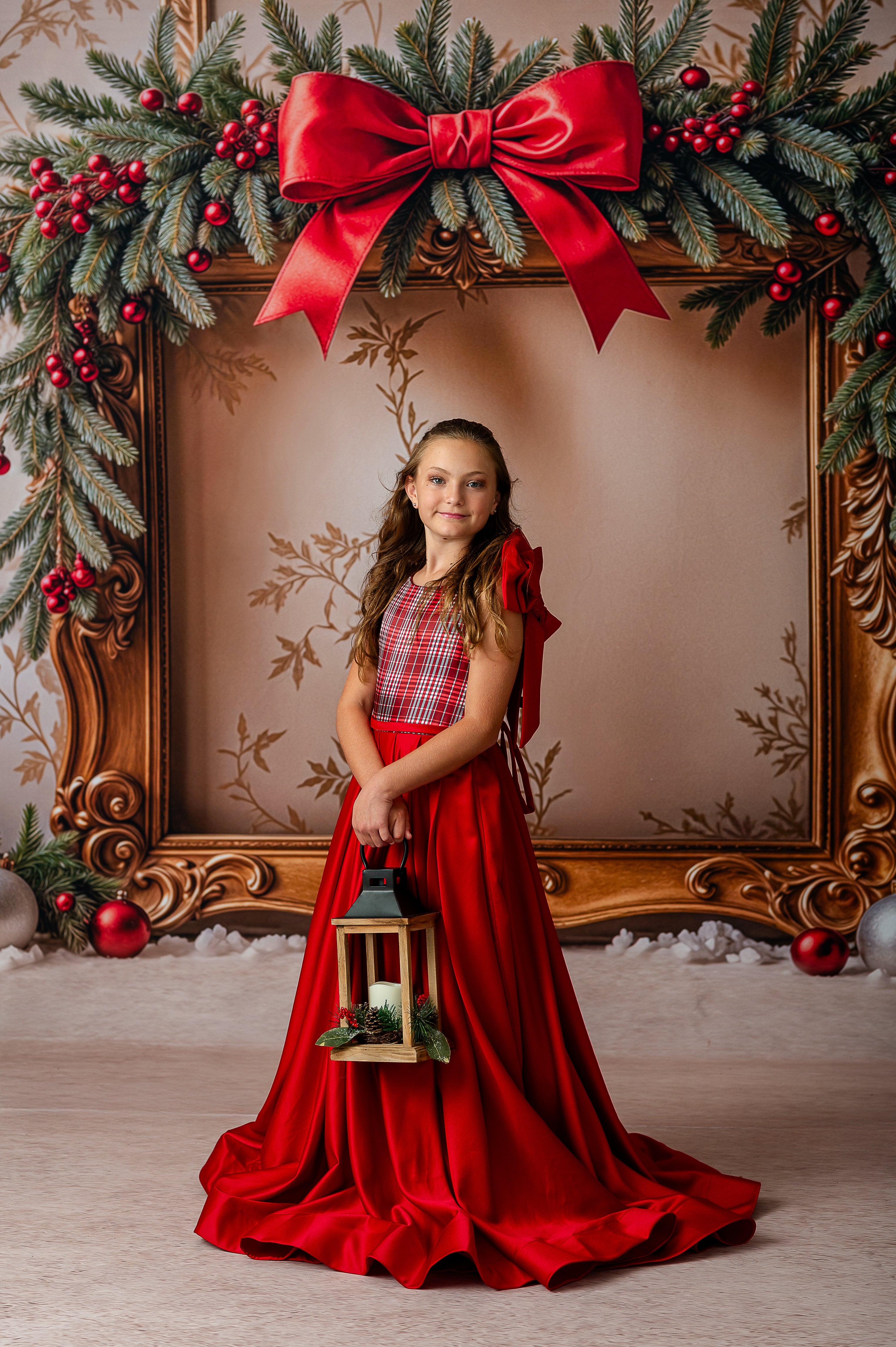 Young girl in a red dress holding a lantern, standing in front of a decorated Christmas frame.