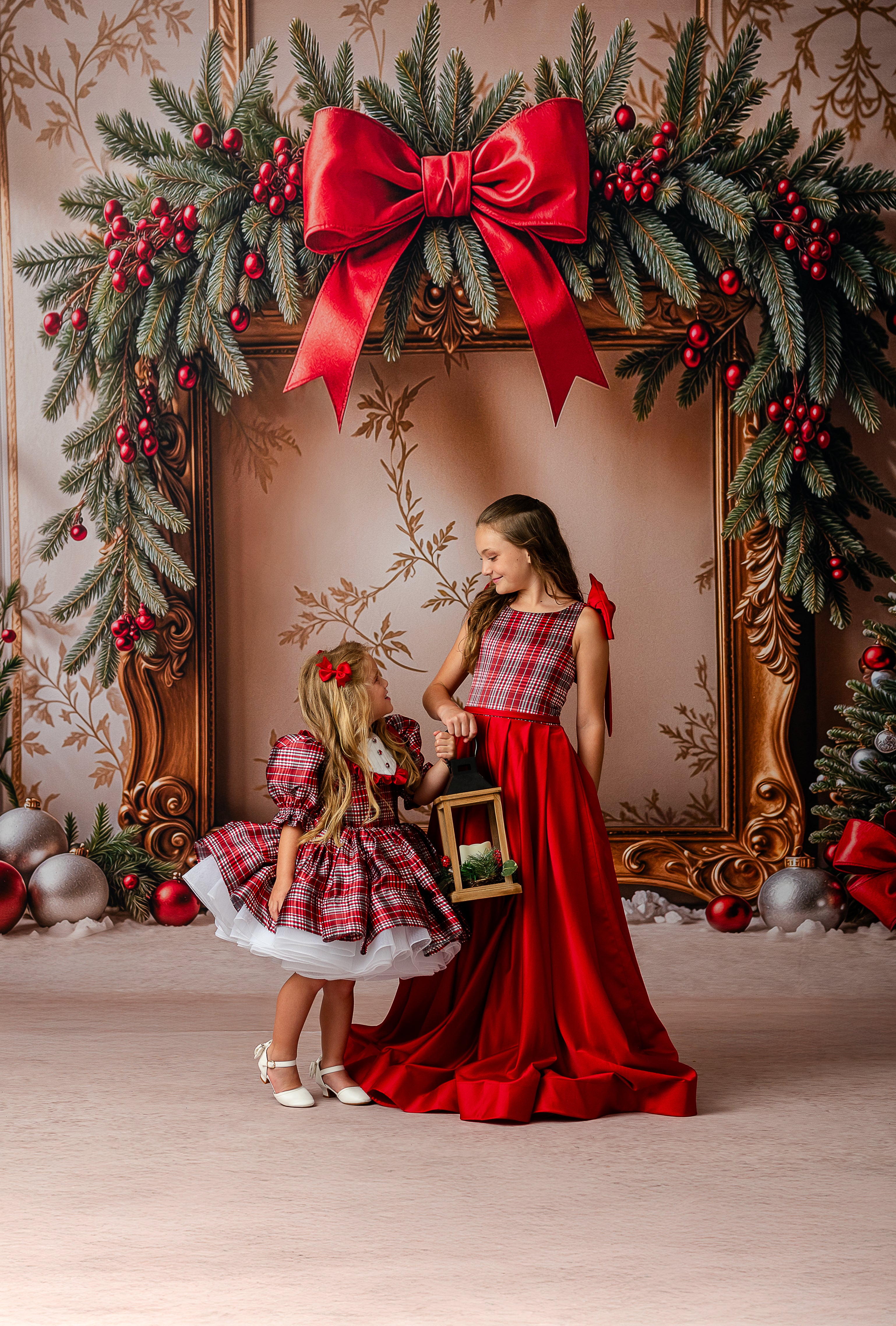 Two children in matching red and plaid dresses standing in front of a decorated Christmas frame.