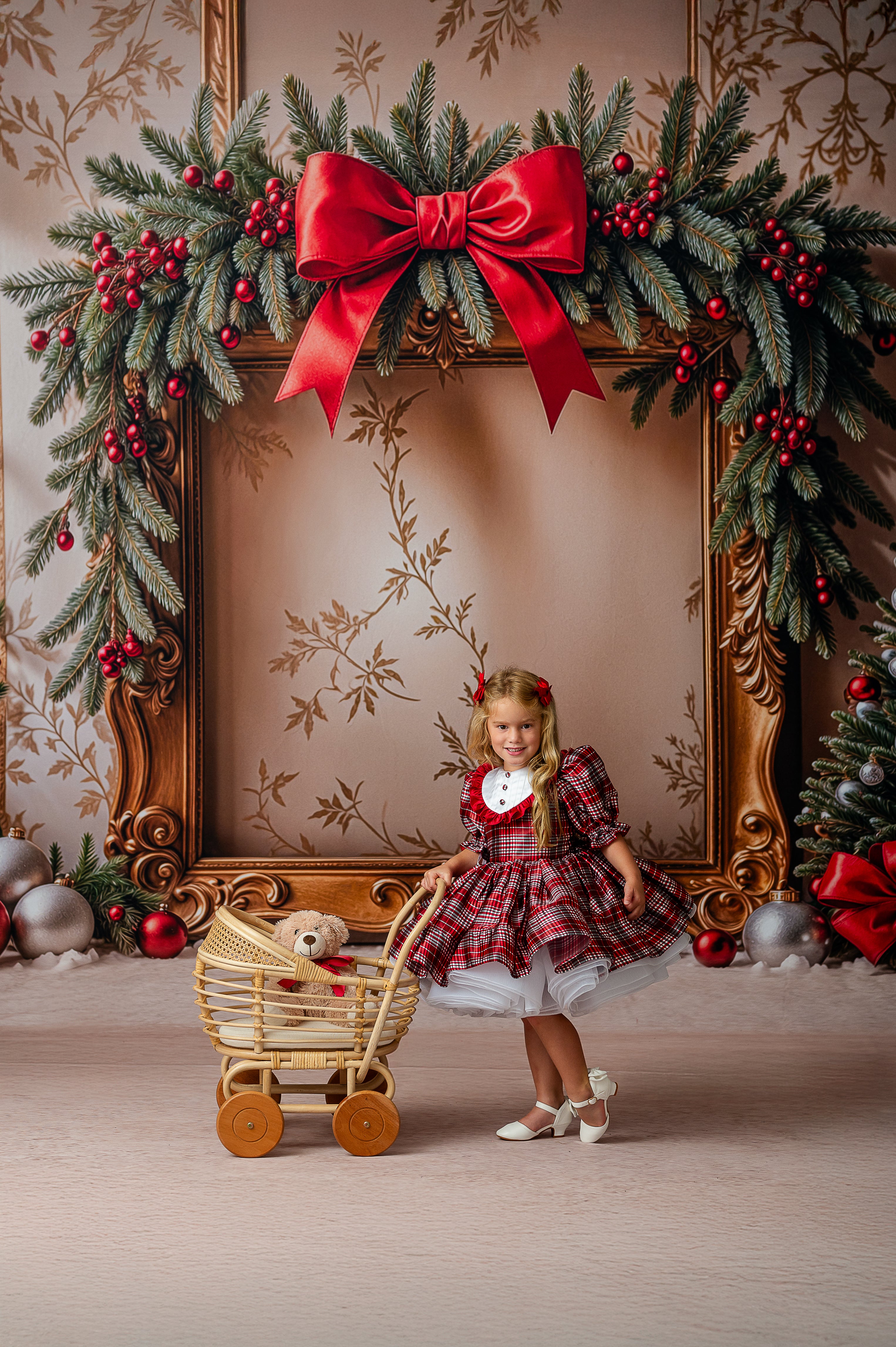Young girl in a plaid dress standing next to a wooden toy cart with a teddy bear, in front of a decorated Christmas frame.