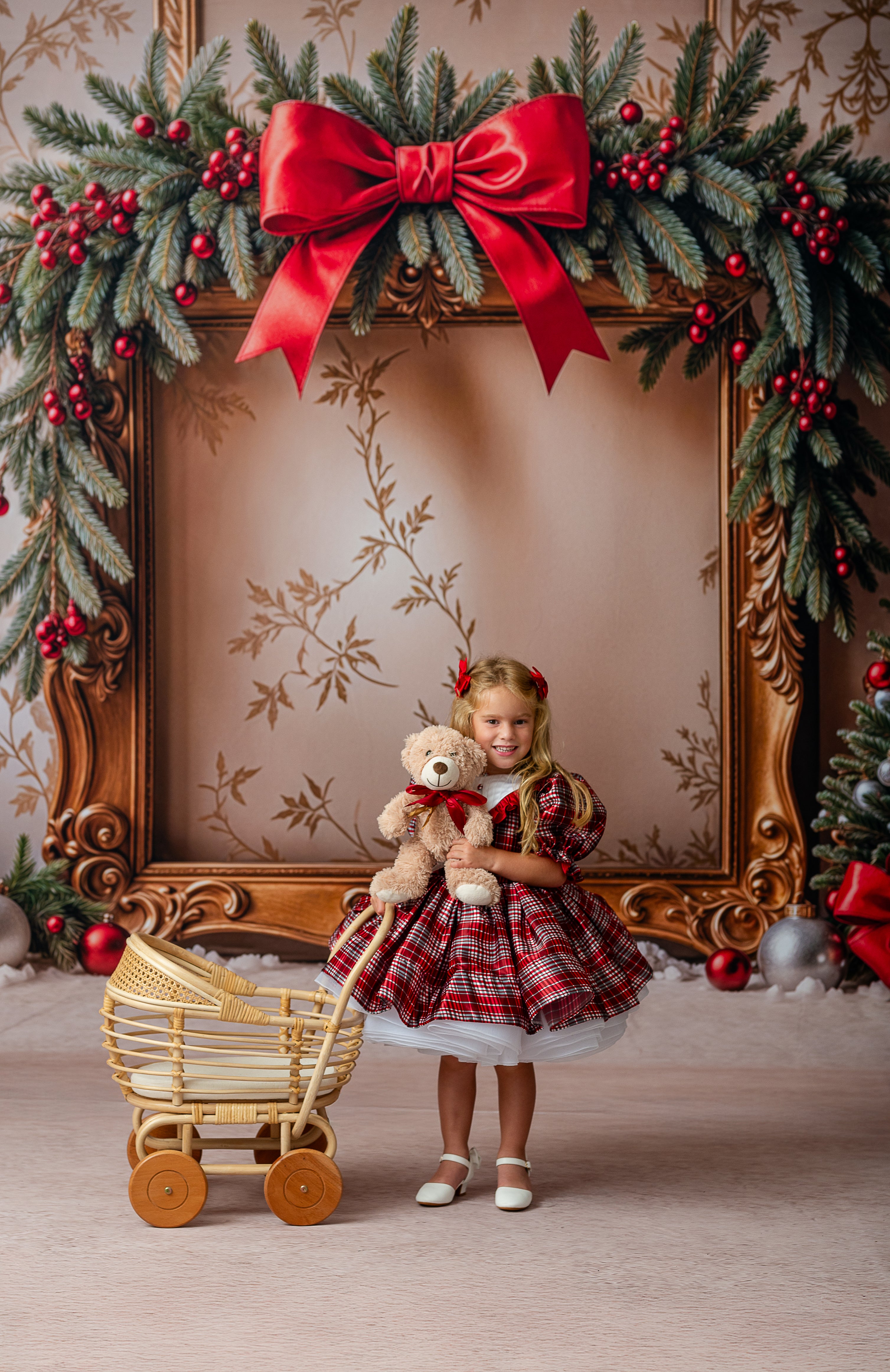 Young girl in a plaid dress holding a teddy bear, standing in front of a decorated Christmas frame.