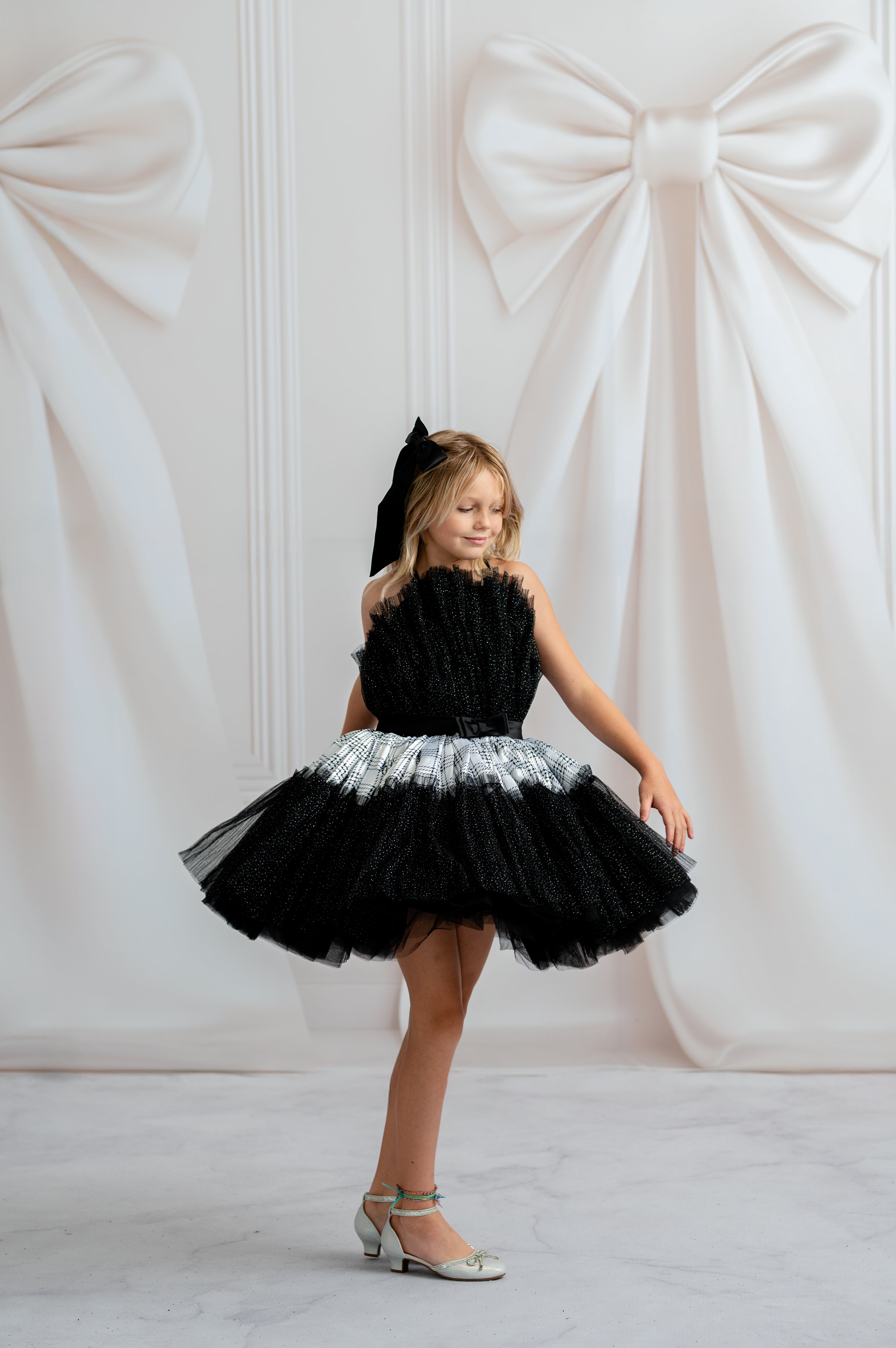 Young girl in a black and silver dress standing against a white wall with decorative bows.