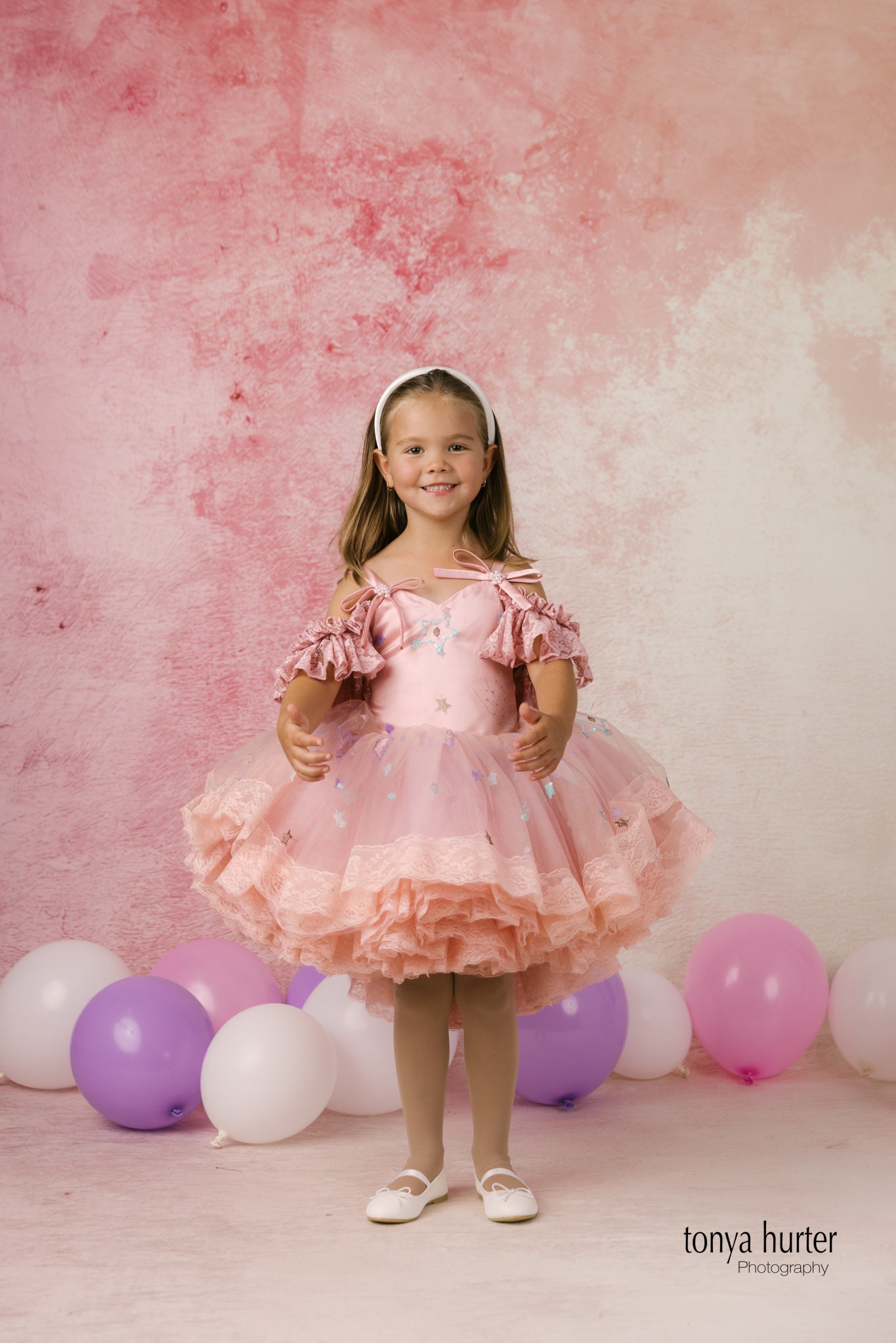 Young girl in a pink dress standing in front of balloons with a pink and white background.