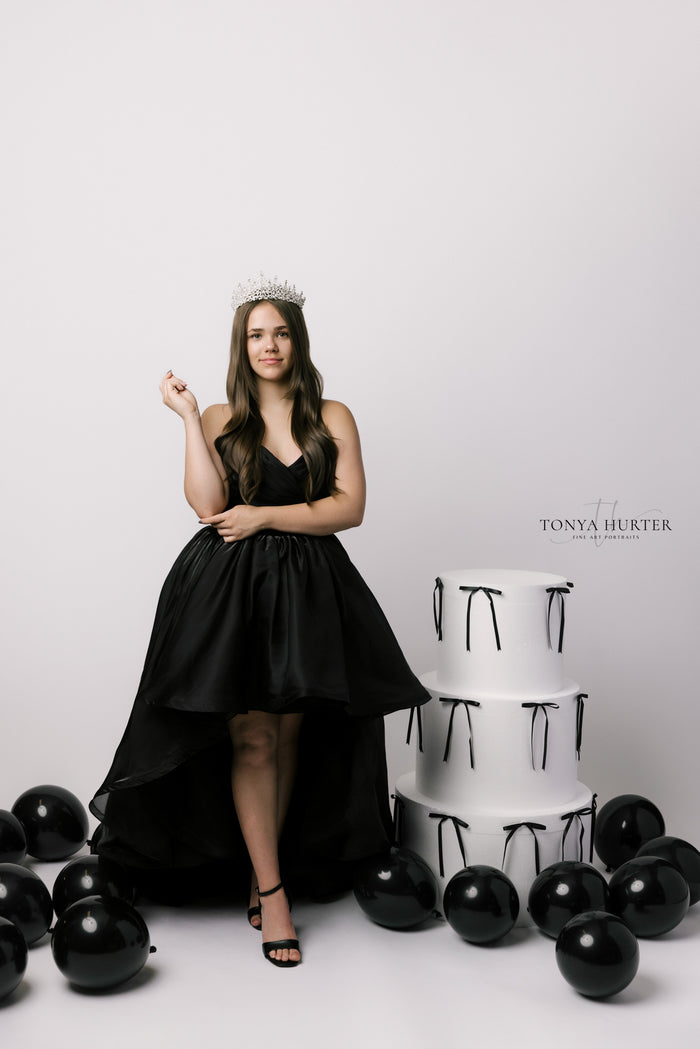 Woman in a black dress standing next to a white cake with black ribbons on a white background.