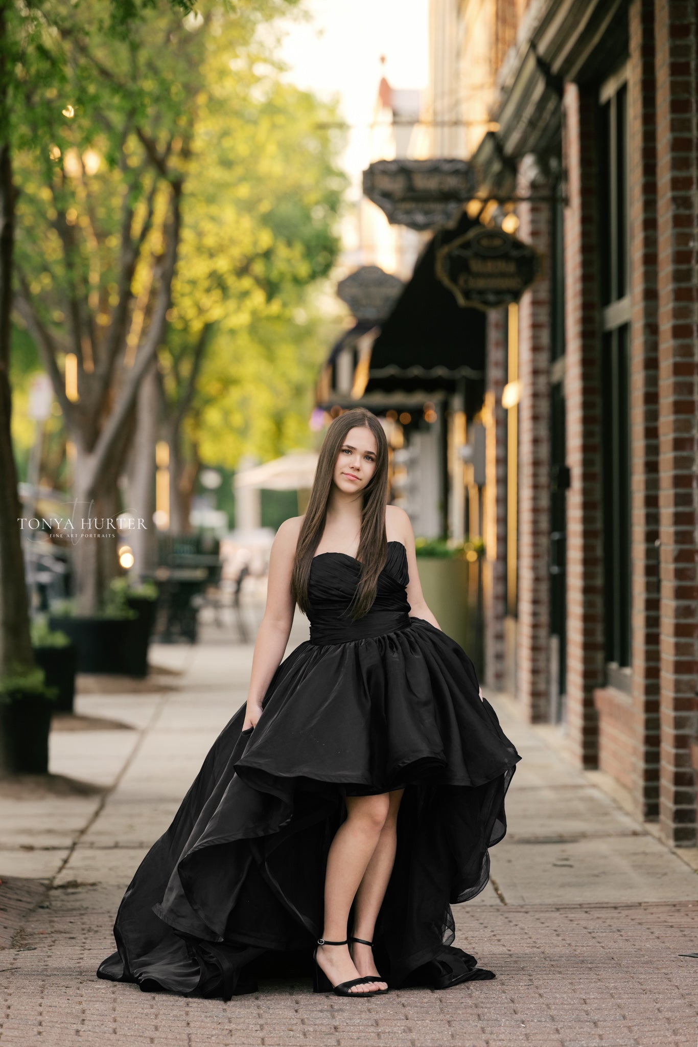 Woman in a black dress standing on a city street with buildings and trees in the background.
