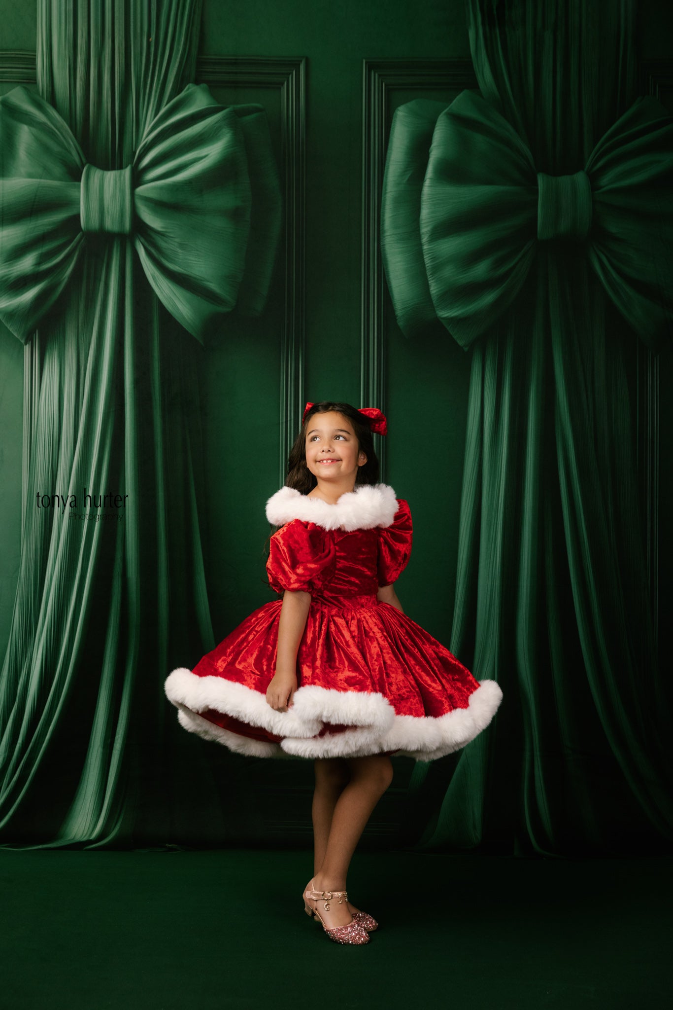 Young girl in a red dress with white fur trim standing in front of green curtains with large bows.