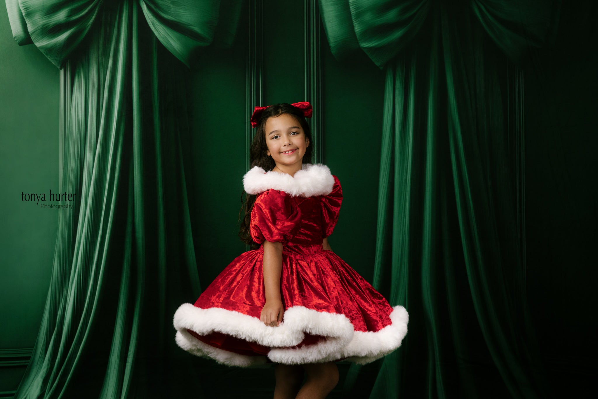 Young girl in a red dress with white fur trim sitting against a green curtain backdrop.