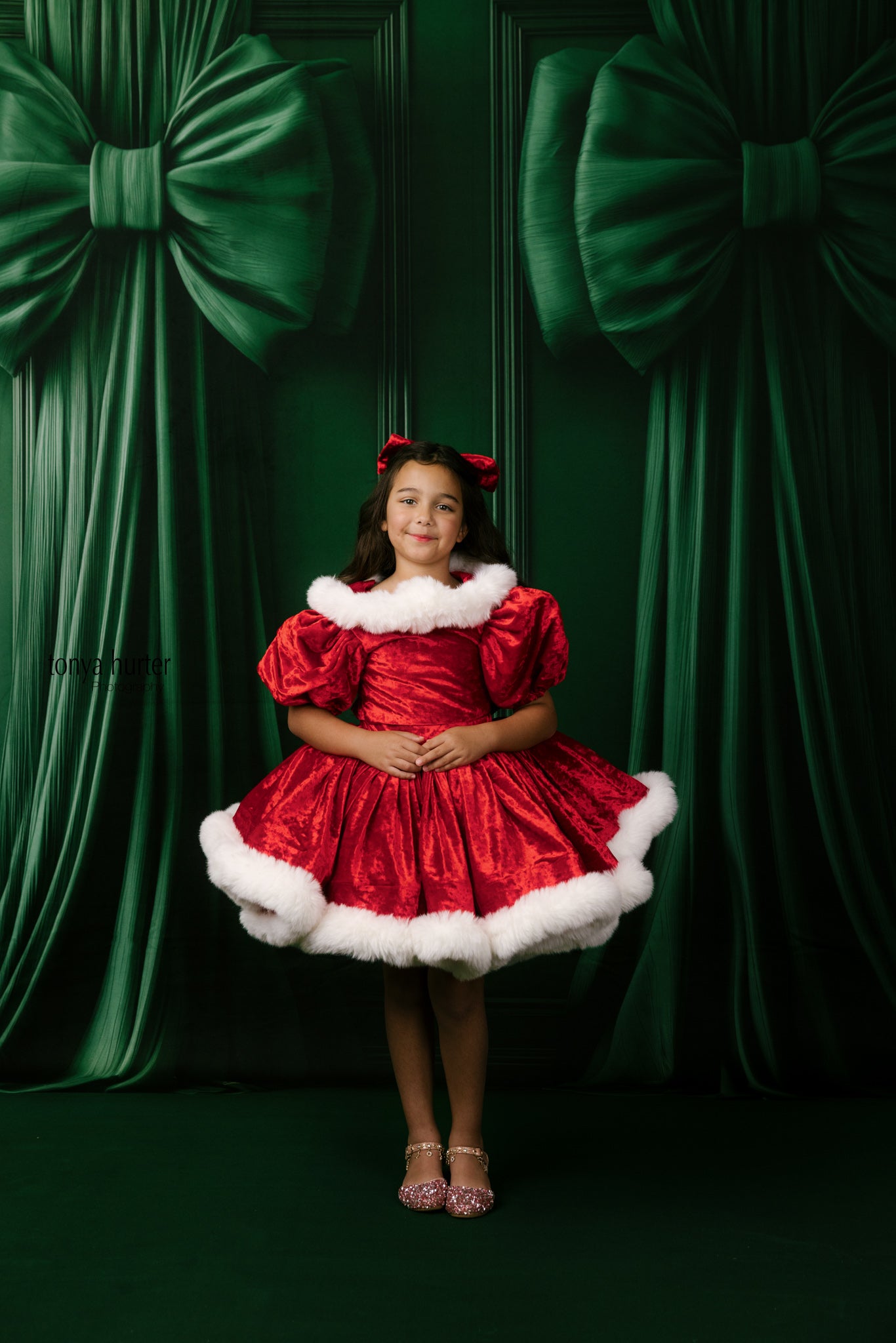 Young girl in a red dress with white fur trim standing in front of green curtains with large bows.