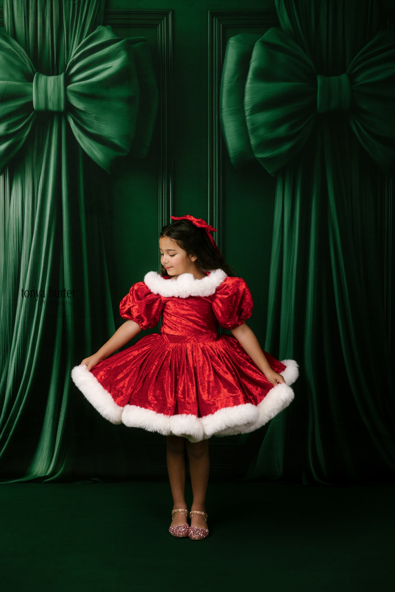 Young girl in a red dress with white fur trim standing against a green curtain backdrop.