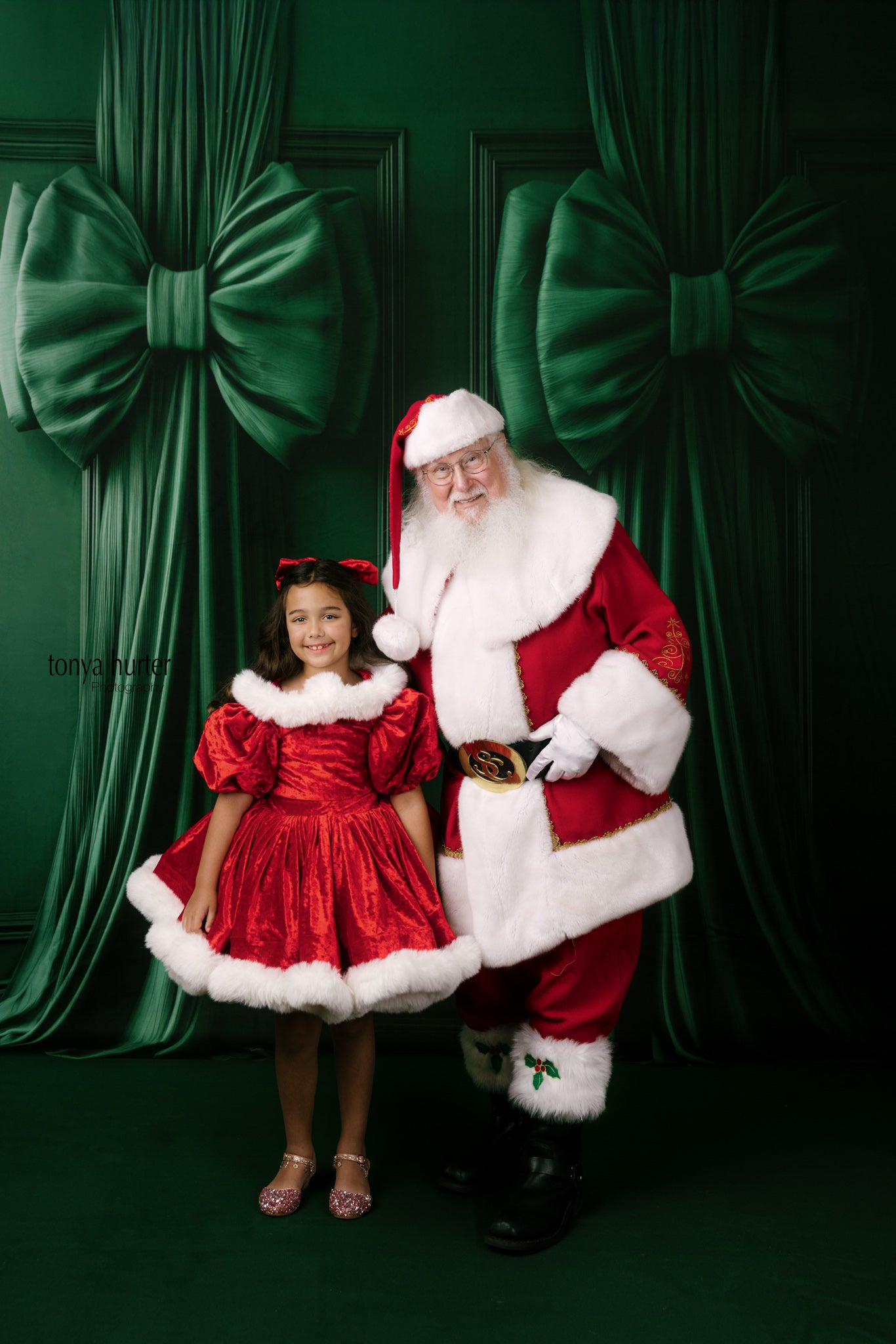 Santa Claus and a young girl in a red dress standing in front of green curtains with large bows.