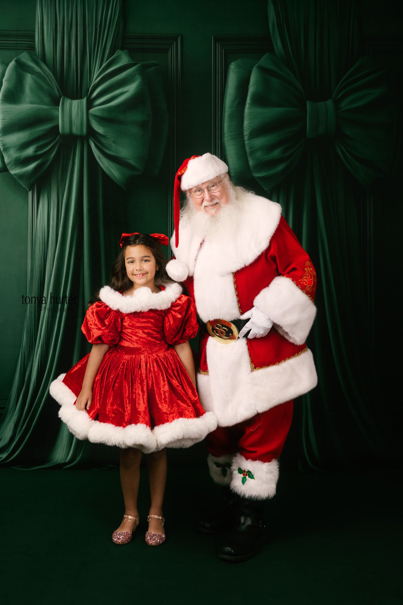 Santa Claus and a young girl in red outfits posing in front of a green curtain with large bows.