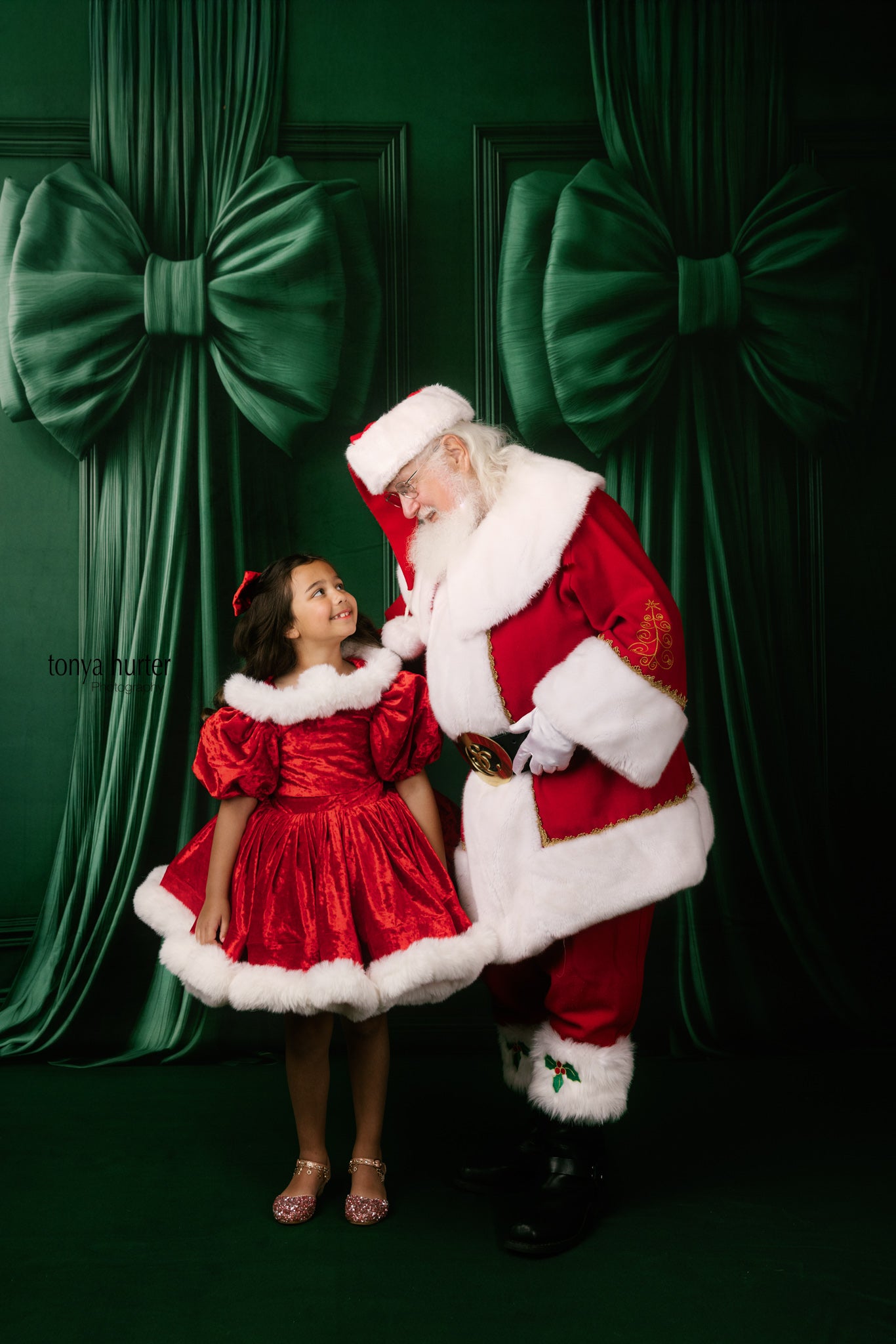 Santa Claus and a young girl in red dresses standing in front of green curtains with large bows.