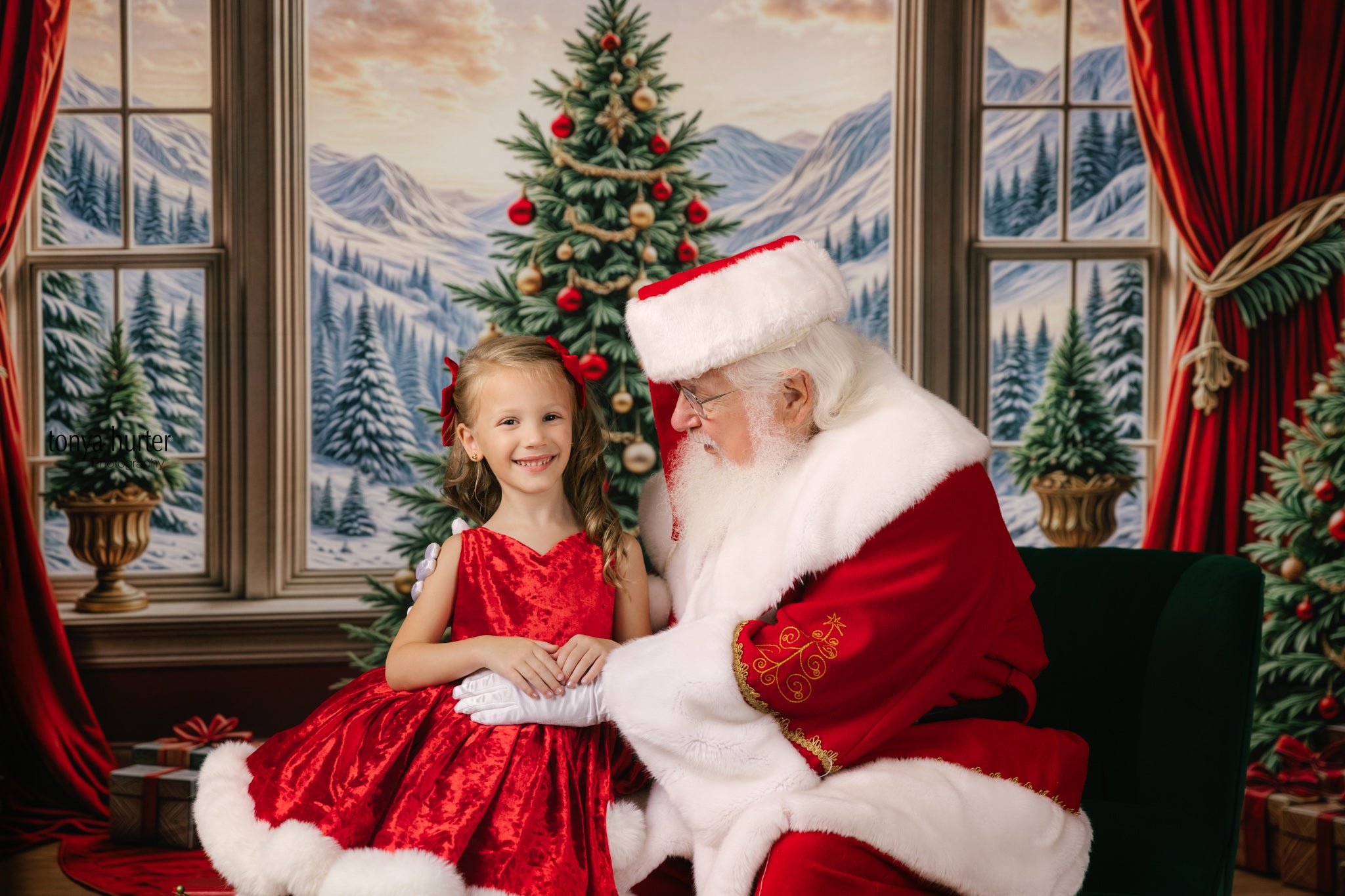 Santa Claus sitting with a young girl in a red dress in a festive setting with Christmas trees and decorations.