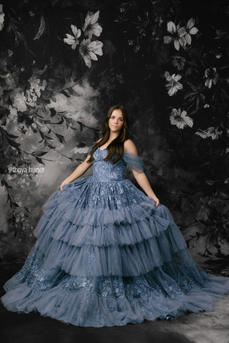 Woman in a blue gown against a dark floral backdrop